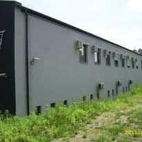 Szary budynek z rzędem klimatyzatorów pod oknami, otoczony zielenią. Widoczne liczne jednostki zewnętrzne klimatyzacji na elewacji budynku.