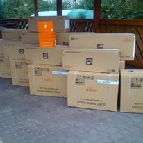 Stos kartonowych pudeł z logo Fujitsu General Limited, prawdopodobnie klimatyzatorów, ułożonych na zewnątrz pod zadaszeniem. Widoczne detale opakowań i otoczenie.