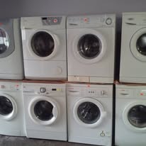 Rząd używanych, białych pralek różnych marek, ustawionych obok siebie, z widocznymi drzwiczkami i panelami sterowania, sugerujący komis lub magazyn sprzętu AGD.