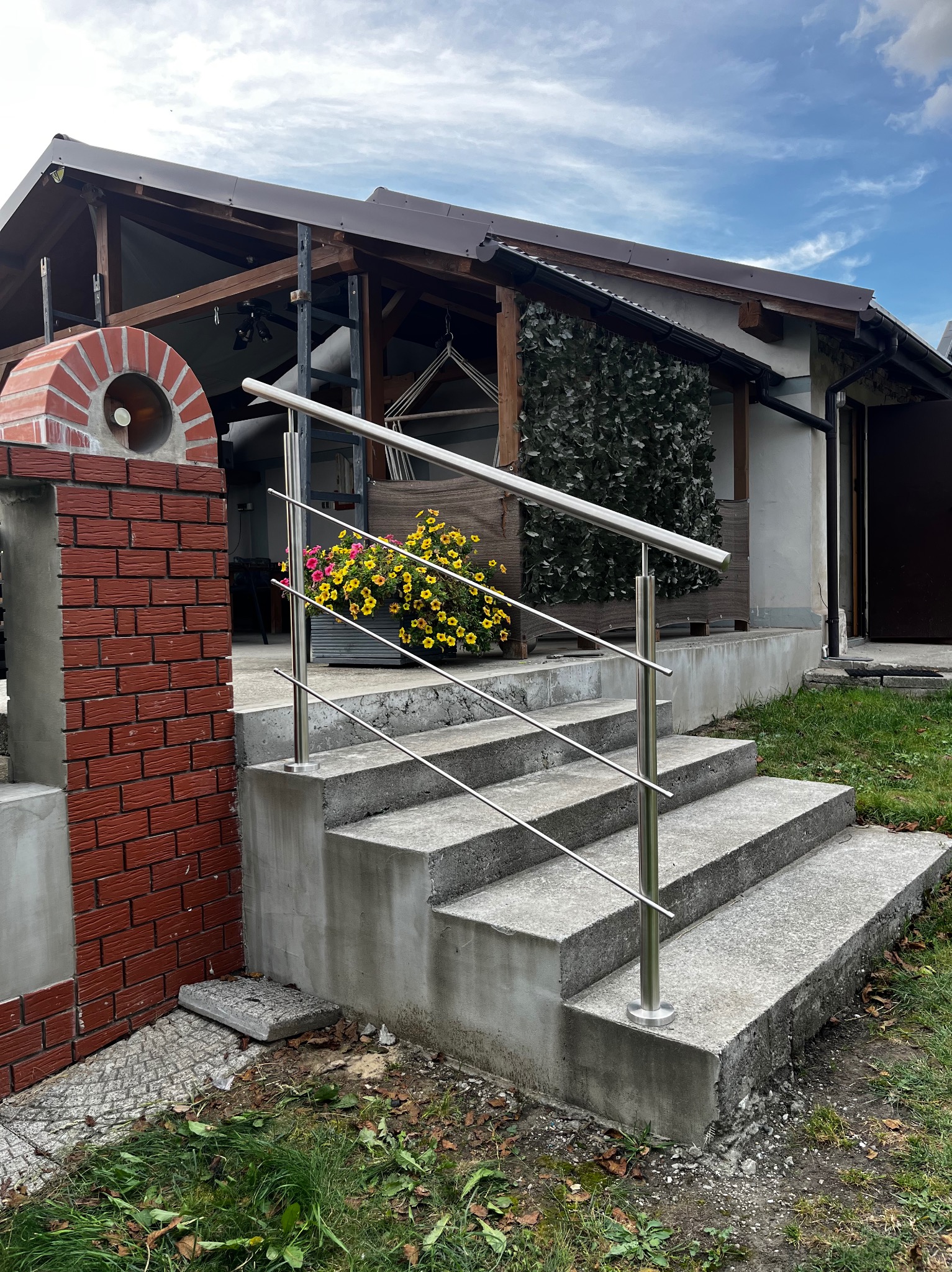 Zewnętrzne schody betonowe z nowoczesną, prostą balustradą ze stali nierdzewnej. Na schodach donica z kwiatami. W tle budynek z elementami drewnianymi.