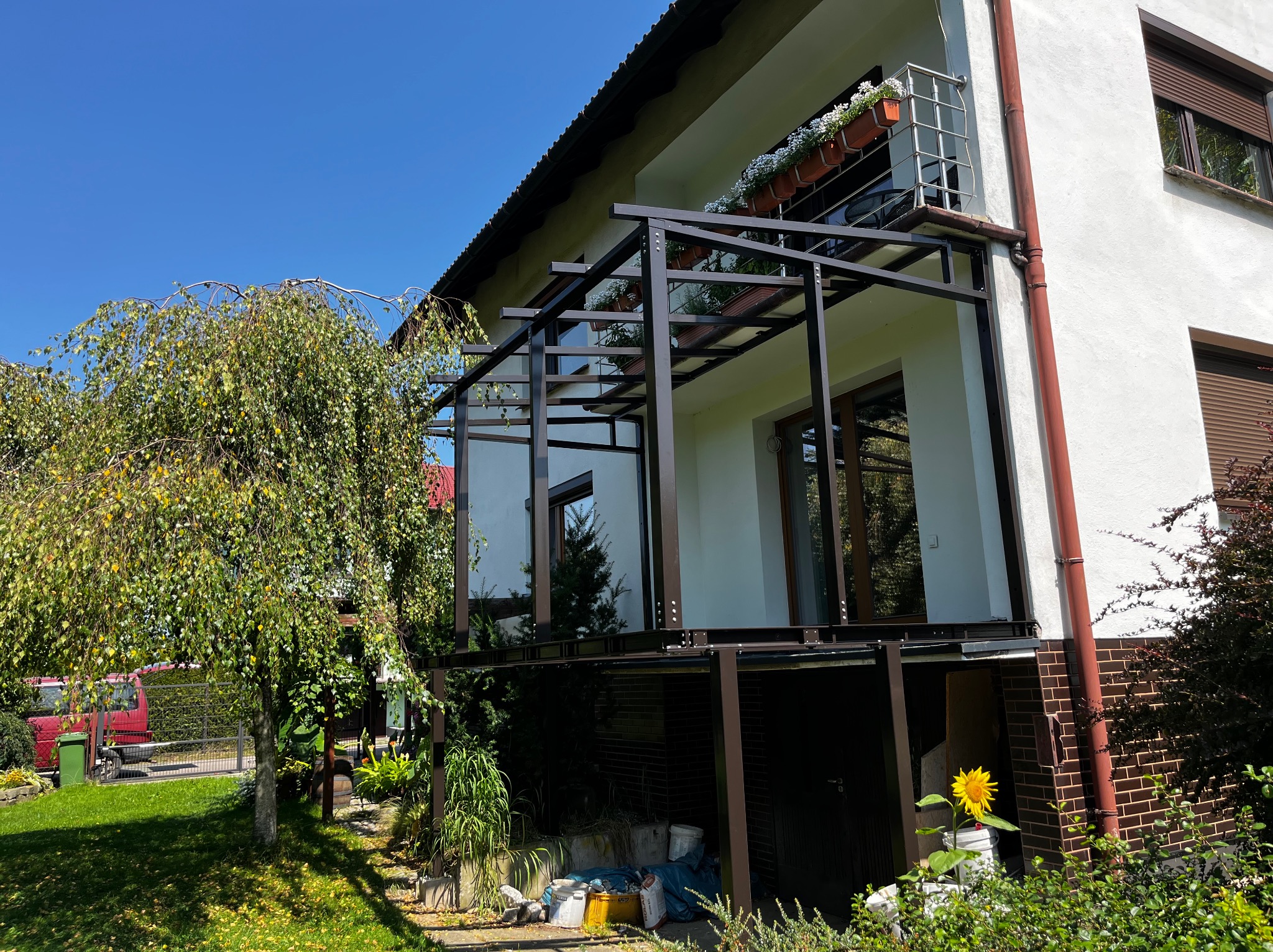 Konstrukcja metalowa pergoli przy domu z balkonem, widok z zewnątrz, w tle zieleń ogrodu i błękitne niebo. Elementy mocowane na śruby.