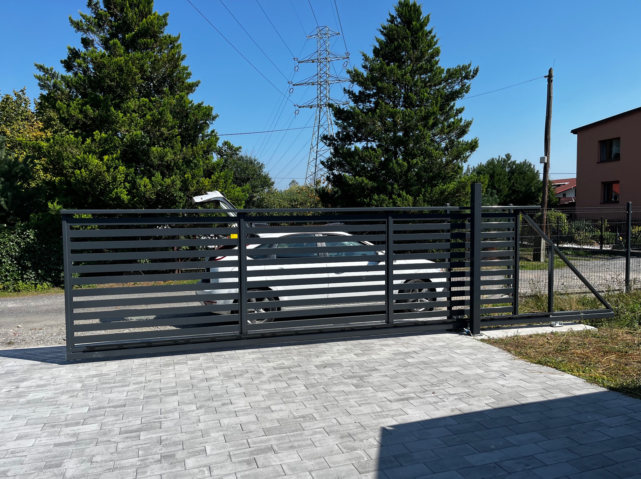 Nowoczesna, przesuwna brama wjazdowa z poziomymi profilami, szara, widoczna na tle brukowanej nawierzchni i zieleni. W tle słup wysokiego napięcia.