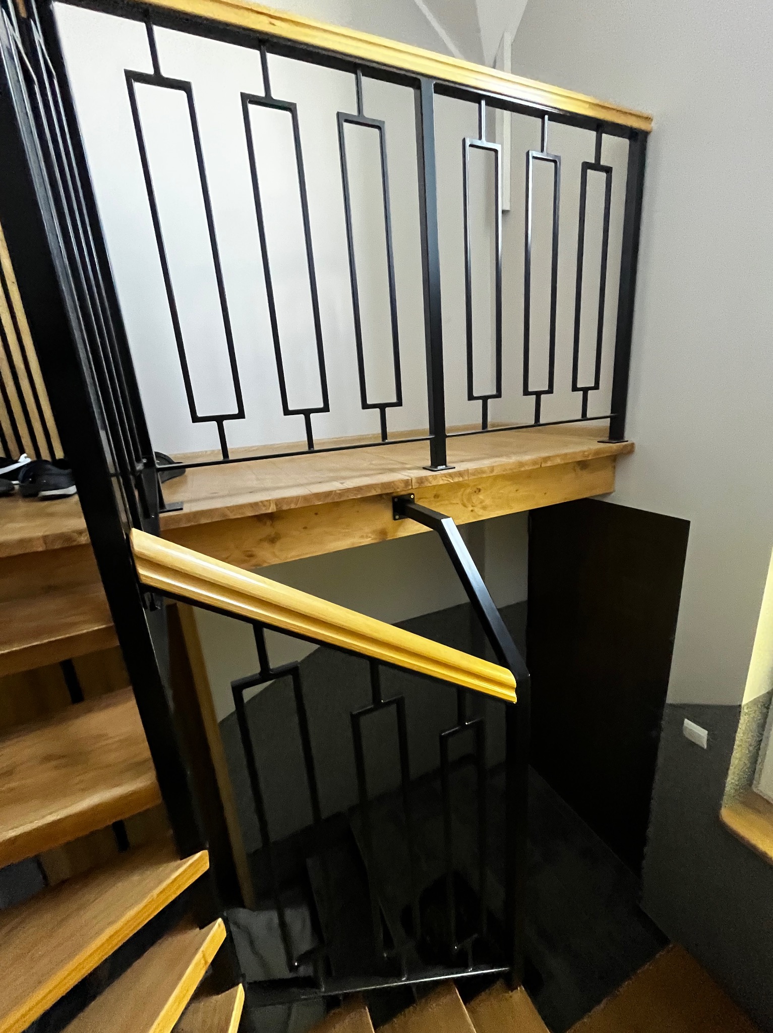 Nowoczesne schody z drewnianymi stopniami i czarną metalową balustradą o geometrycznym wzorze, wykończoną jasnym drewnem. Widok z góry na dół.