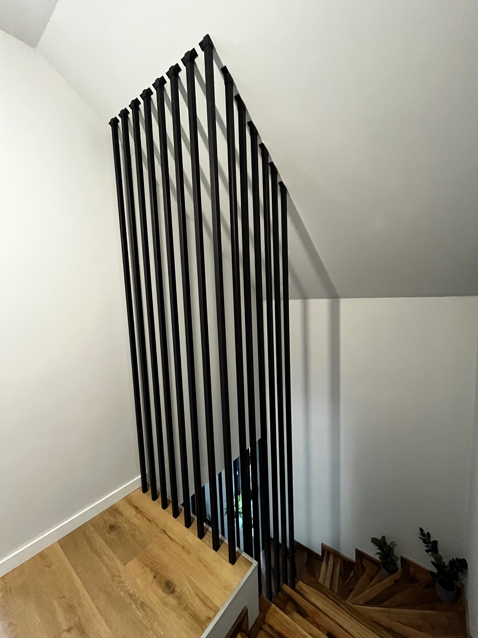 Nowoczesna balustrada schodowa z czarnych, pionowych profili, drewniane schody, białe ściany. Minimalistyczny design z roślinami w tle. Ujęcie z góry.