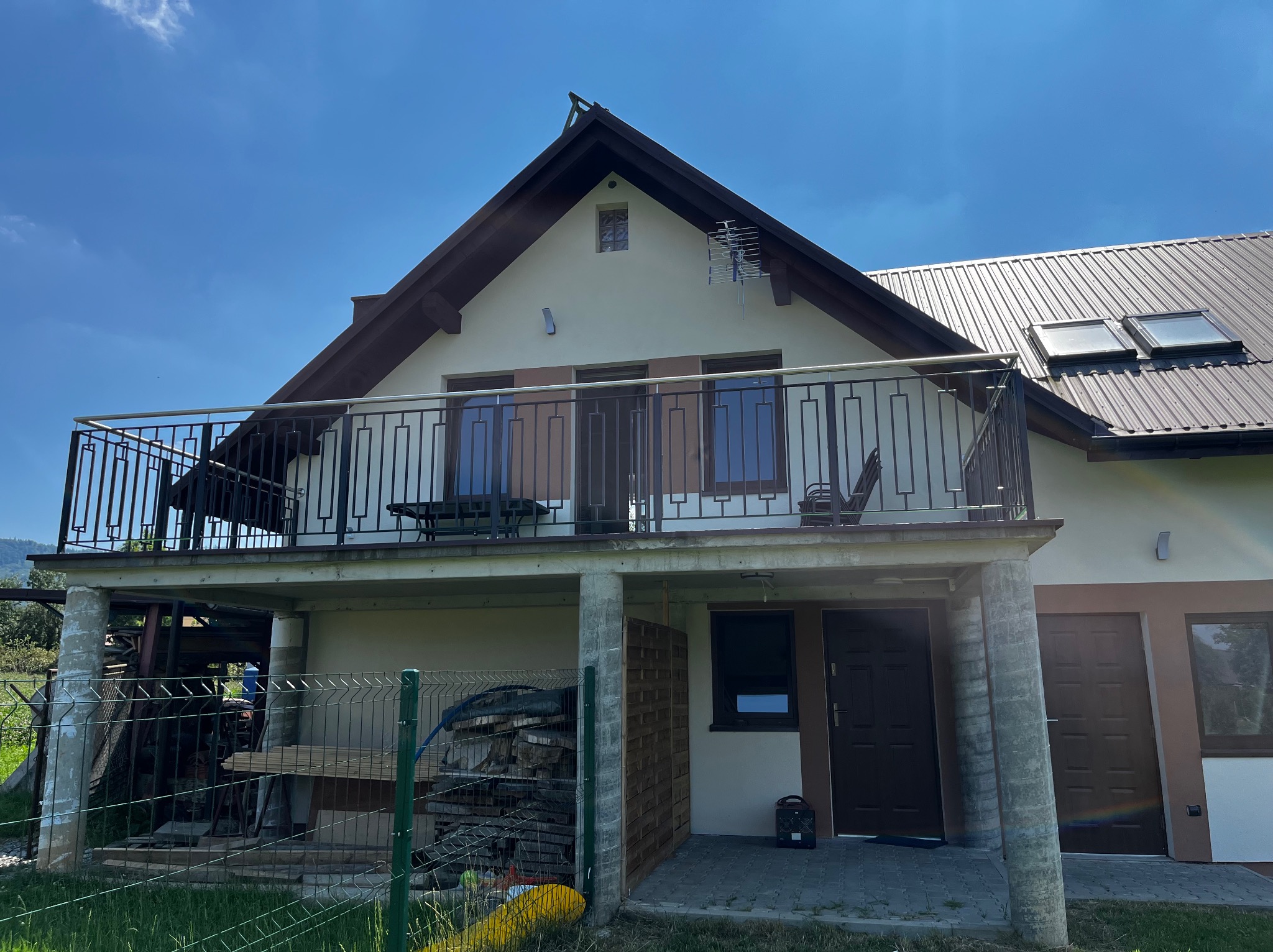 Dom z balkonem zabezpieczonym czarną, metalową balustradą o nowoczesnym designie. Widoczne elementy konstrukcyjne i otoczenie budynku.