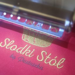 LEDA SPORT JOANNA SOWA - Czerwona tkanina z wyhaftowanym złotym napisem 'Słodki Stół by Pastuszka' pod ramką maszyny do haftowania, ukazująca detale i precyzję wykonania.