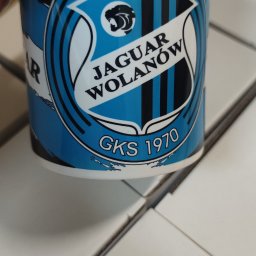 LEDA SPORT JOANNA SOWA - Biały kubek z niebiesko-czarnym nadrukiem herbu klubu sportowego 'Jaguar Wolanów GKS 1970', trzymany w dłoni, na tle białych pudełek.