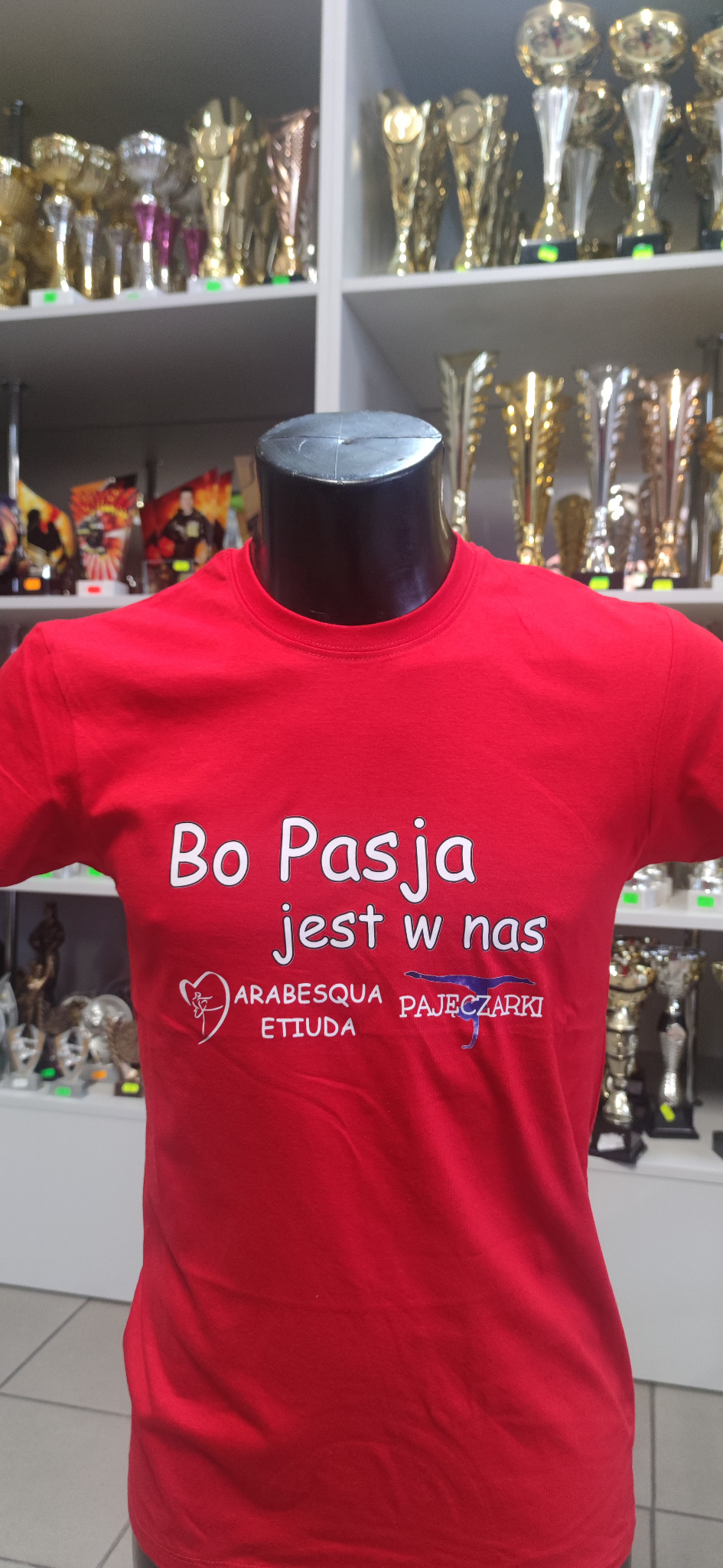 Czerwona koszulka z białym nadrukiem 'Bo Pasja jest w nas' oraz logotypami 'Arabesqua Etiuda' i 'Pajęczarki', prezentowana na manekinie na tle półek z trofeami.