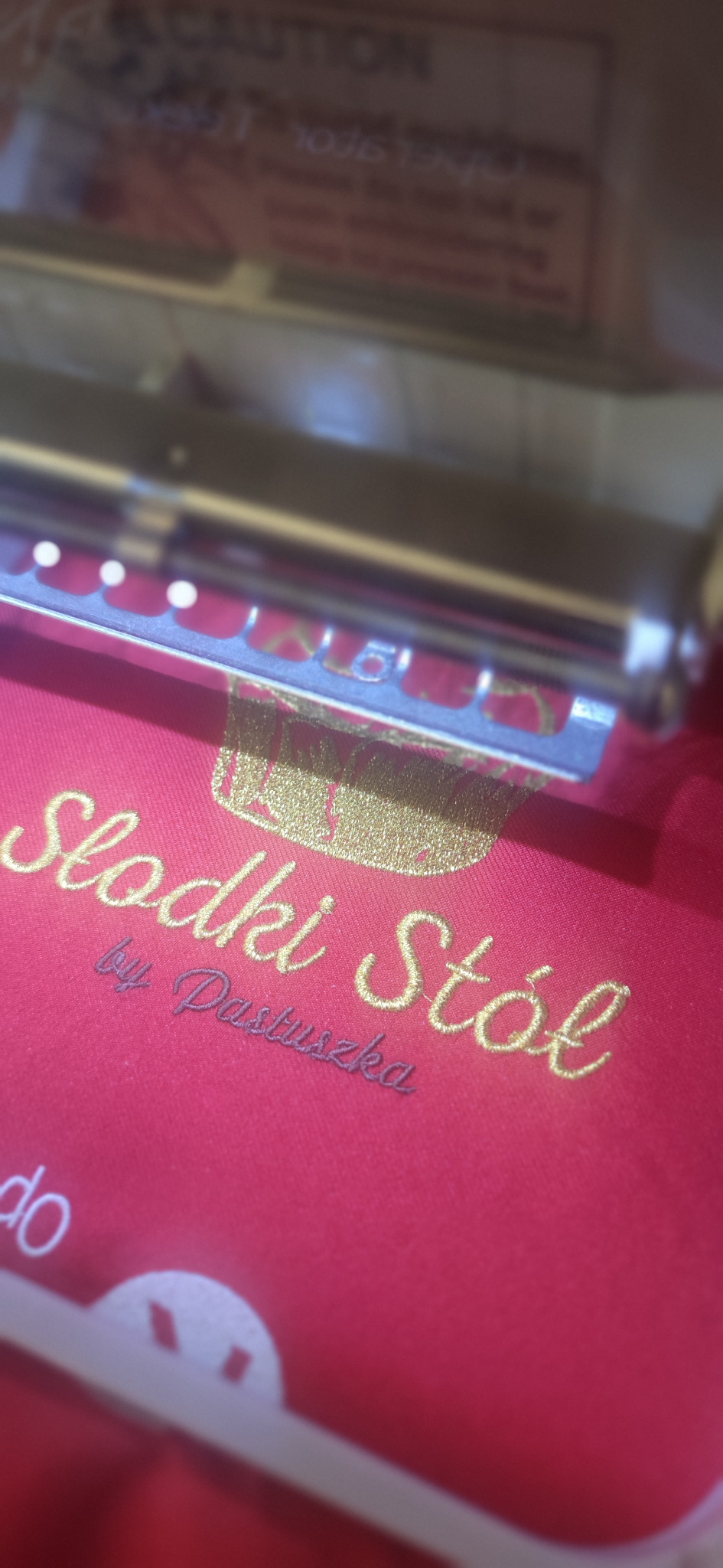 Czerwona tkanina z wyhaftowanym złotym napisem 'Słodki Stół by Pastuszka' pod ramką maszyny do haftowania, ukazująca detale i precyzję wykonania.