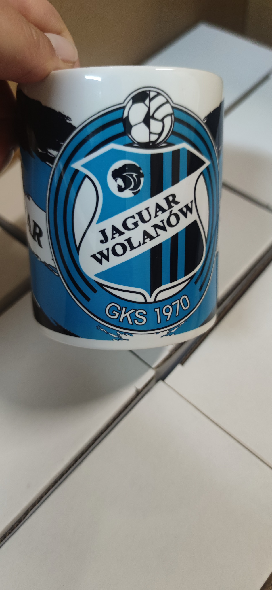 Biały kubek z niebiesko-czarnym nadrukiem herbu klubu sportowego 'Jaguar Wolanów GKS 1970', trzymany w dłoni, na tle białych pudełek.