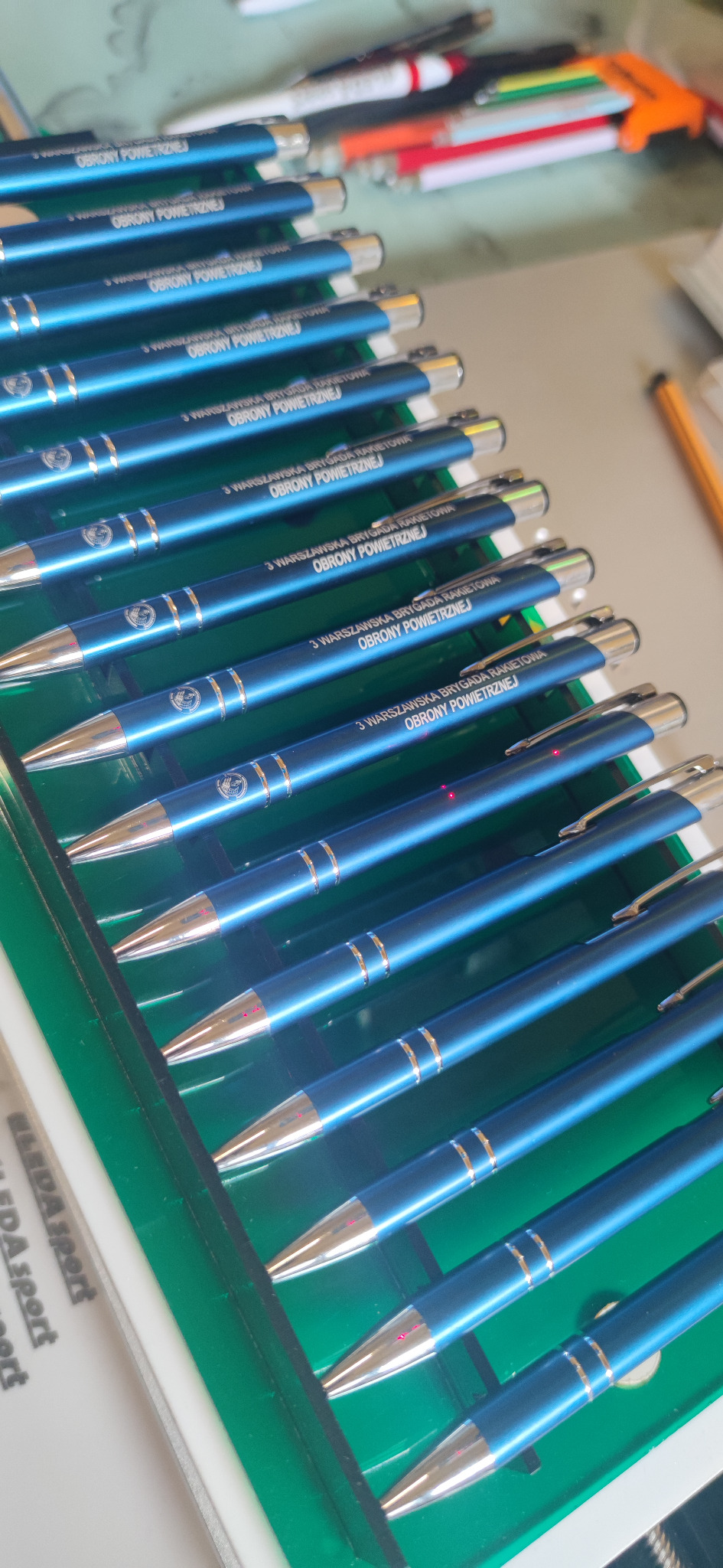 Zestaw niebieskich długopisów z nadrukiem '3 Warszawska Brygada Rakietowa Obrony Powietrznej' w zielonym pudełku. Widoczne inne długopisy w tle.
