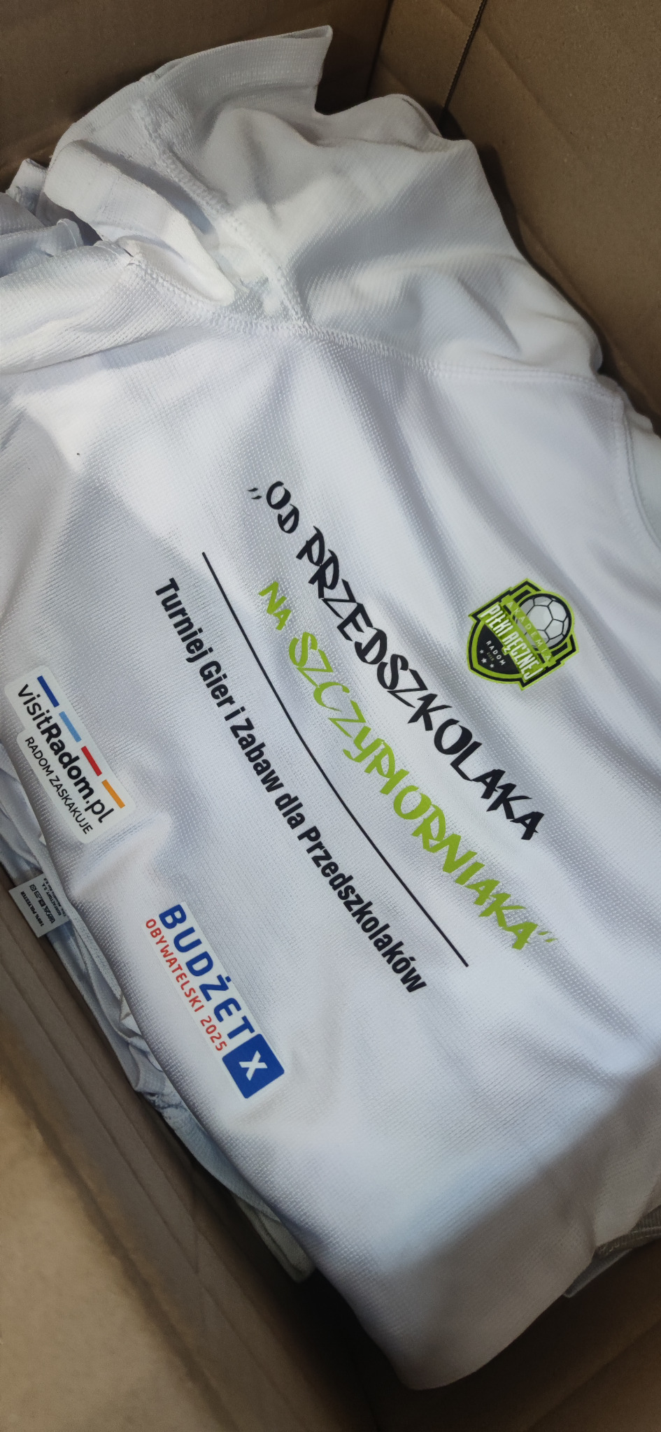 Biała koszulka sportowa z nadrukiem 'Od Przedszkolaka na Szczypiorniaka', logo Akademii Piłki Ręcznej Radom i hasłem 'Budżet Obywatelski 2025' w kartonie.
