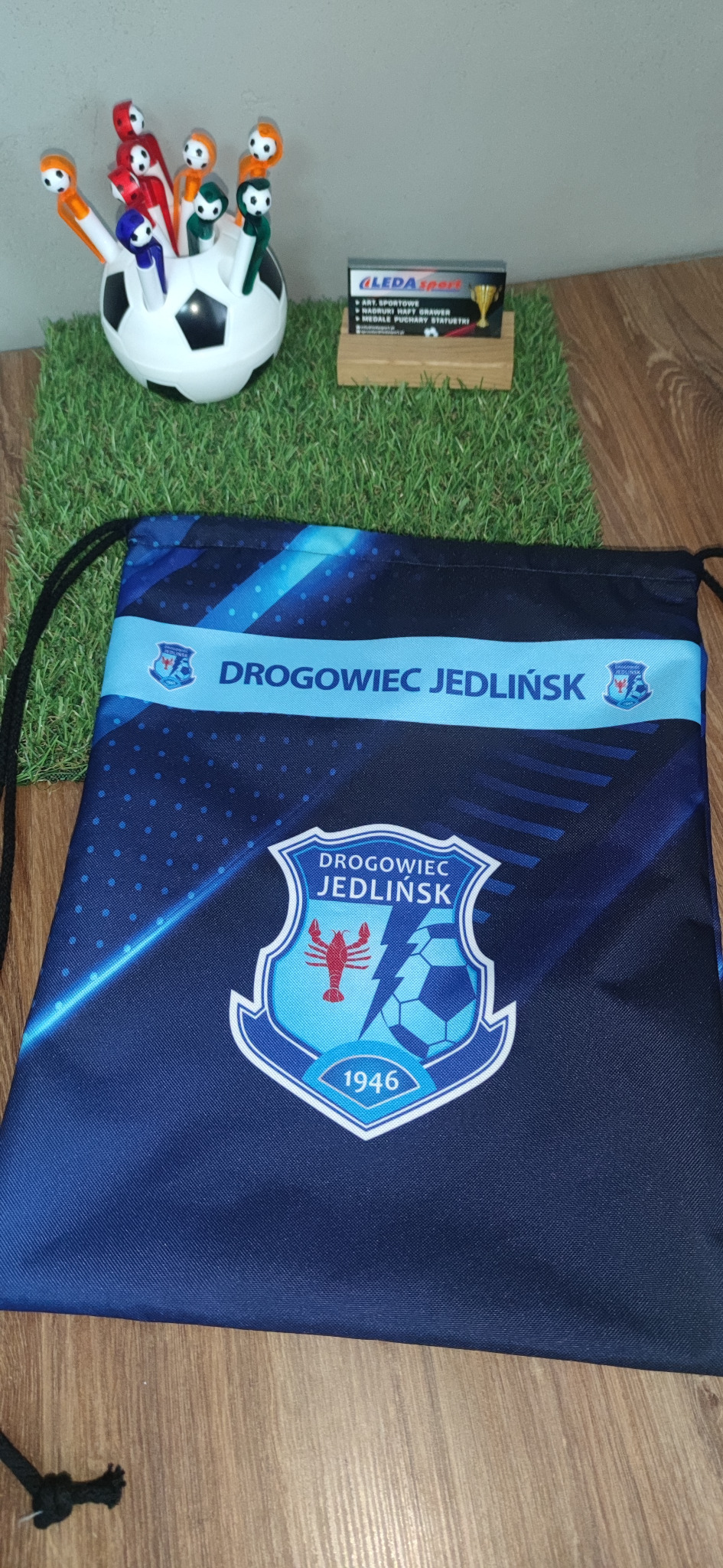 Granatowy worek sportowy z nadrukiem herbu i napisu 'Drogowiec Jedlińsk', leżący na drewnianym blacie. W tle trawa dekoracyjna, piłka z długopisami i wizytówka.