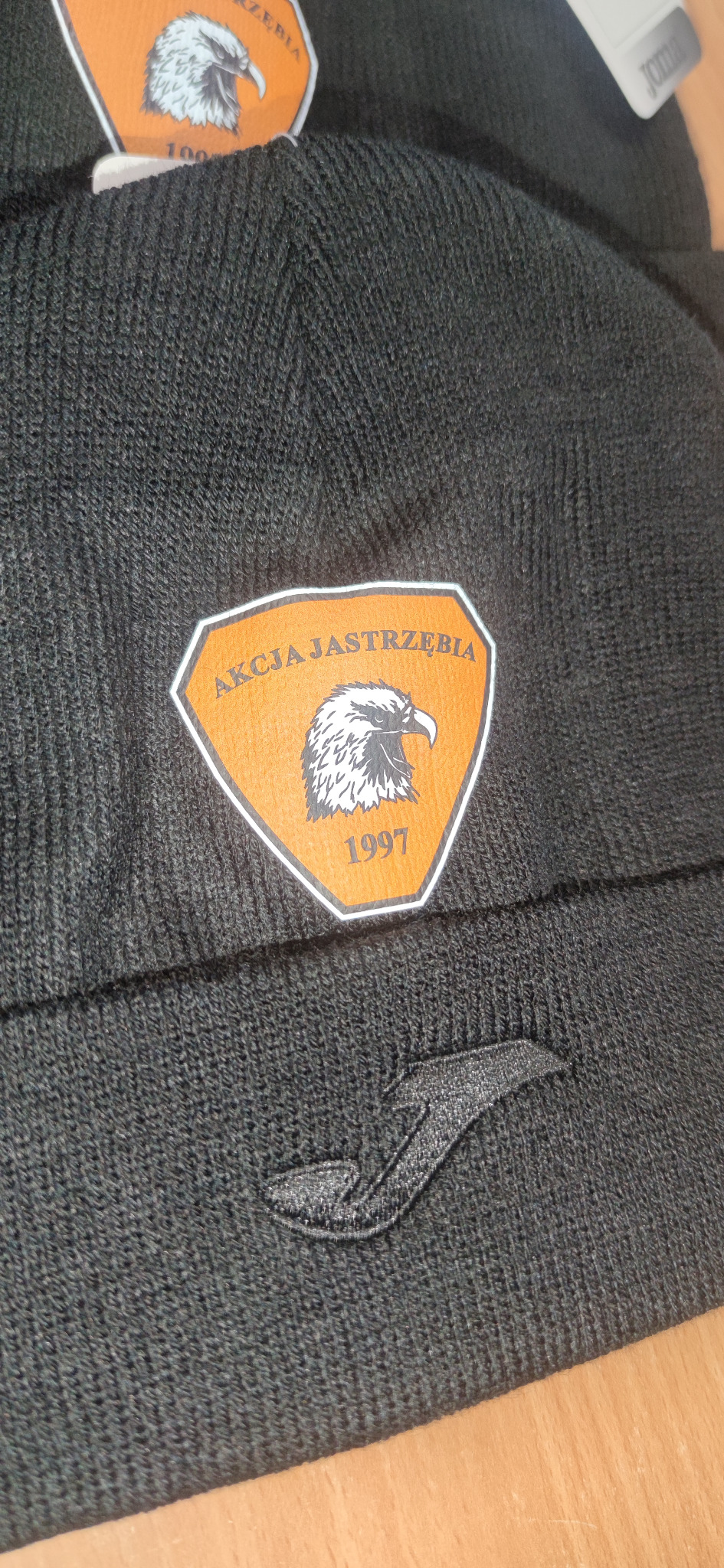Ciemna, dziergana czapka z naszywką 'Akcja Jastrzębia 1997' i logo Joma. Widoczne detale splotu i faktura materiału. Czapka leży na drewnianej powierzchni.