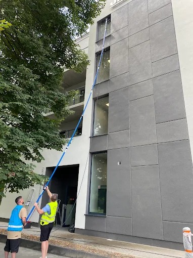 Dwaj pracownicy w kamizelkach czyszczą okna na wysokości za pomocą długiej, niebieskiej lancy. Budynek z szarą elewacją i dużymi oknami. Widoczne drzewo.