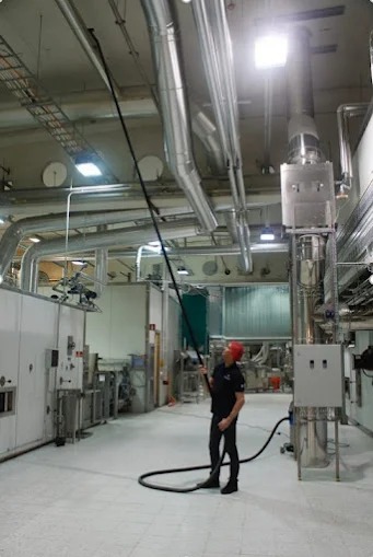 Pracownik w kasku czyści sufit hali przemysłowej za pomocą długiej rury ssącej podłączonej do odkurzacza. Widoczne rury wentylacyjne i instalacje.