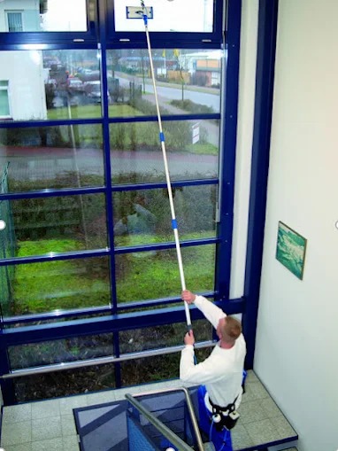 Pracownik w uprzęży czyści okno na wysokości za pomocą długiego kija z myjką. Widok z dołu na okno z niebieską ramą i otoczenie budynku.