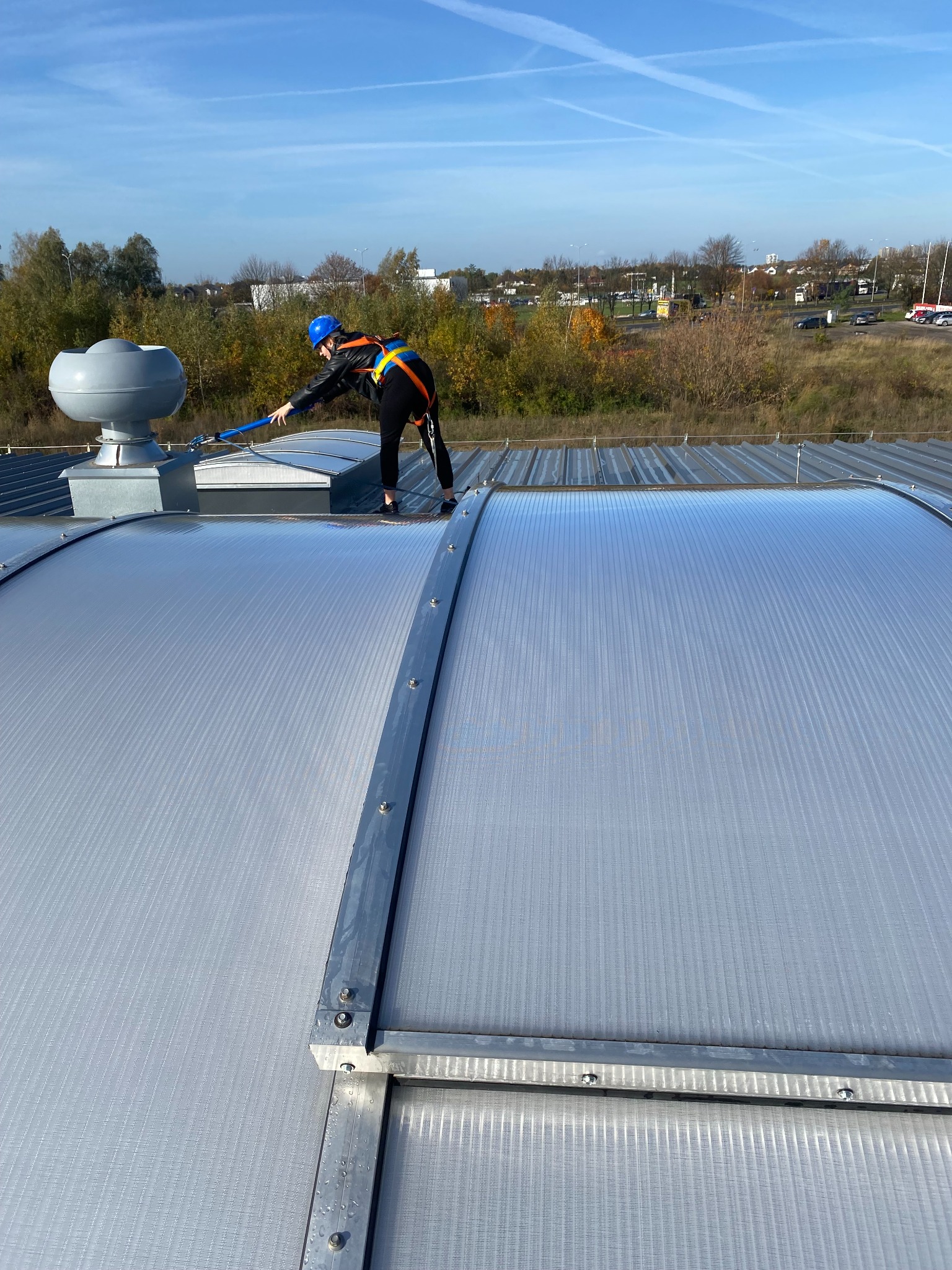 Pracownik w kasku i uprzęży czyści dach przemysłowy za pomocą długiej szczotki. Widoczne elementy konstrukcyjne dachu i otoczenie z zielenią w tle.