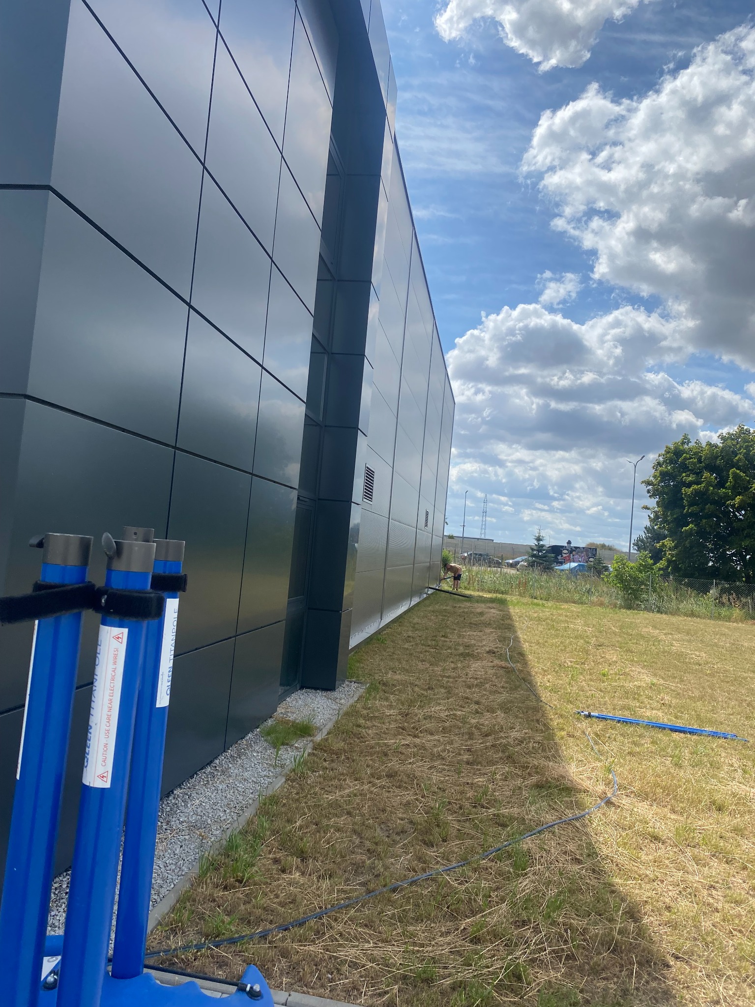 Czyszczenie elewacji budynku komercyjnego z użyciem teleskopowych lanc. Widoczne niebieskie lance i pracownik w oddali. Błękitne niebo z chmurami odbija się w panelach.