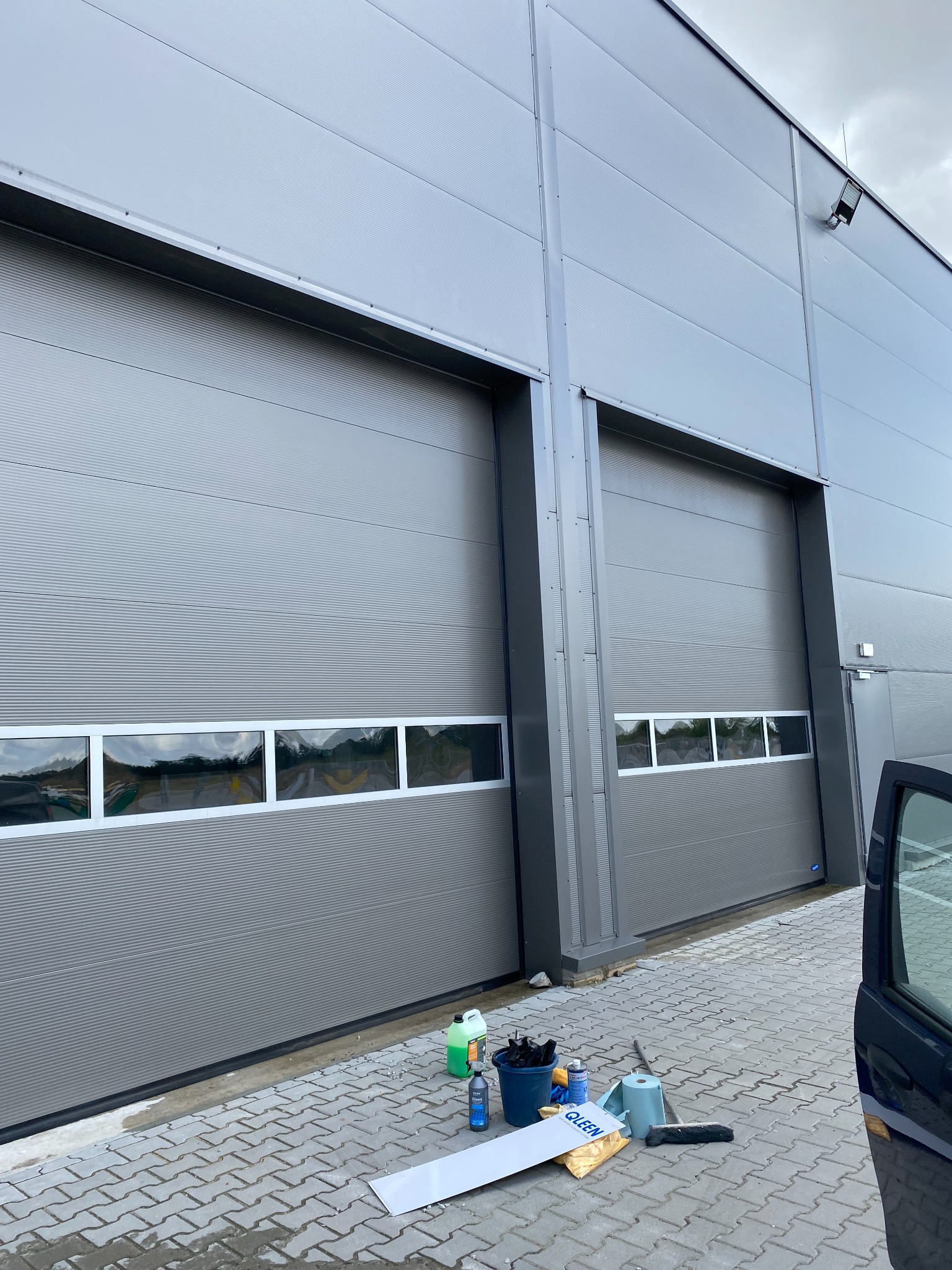 Szare, segmentowe bramy garażowe z oknami po umyciu. Na kostce brukowej wiadro, płyny i akcesoria do mycia. Widoczny fragment otwartych drzwi samochodu.