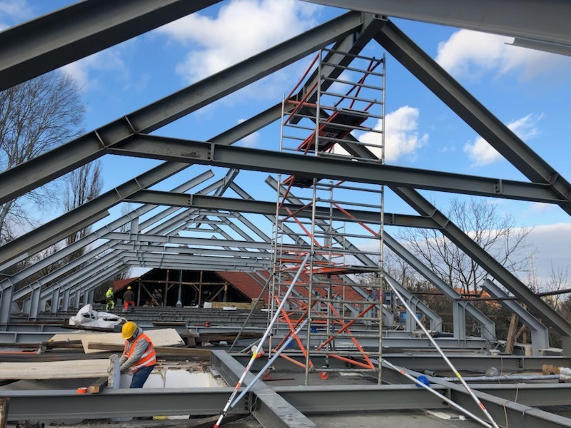 Stalowa konstrukcja dachu w trakcie rozbudowy, widoczny pracownik w kasku i kamizelce, rusztowanie, błękitne niebo w tle. Prace rozbudowy domu w toku.