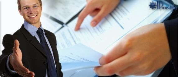 Uśmiechnięty mężczyzna w garniturze wyciąga dłoń na powitanie, w tle dokumenty analizowane przez drugą osobę. Logo firmy w prawym górnym rogu.