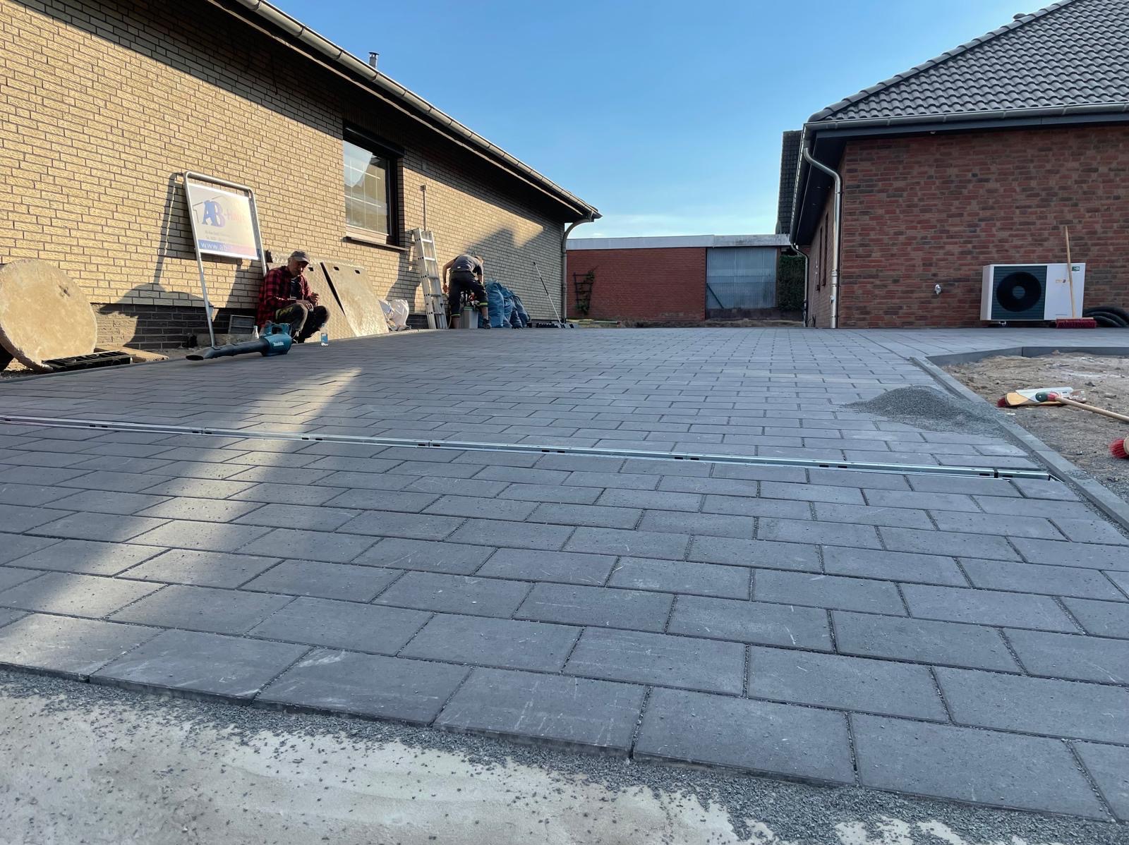 Ułożona, szara kostka brukowa na podjeździe domu jednorodzinnego. W tle pracownicy i sprzęt budowlany. Widoczne detale wykończenia i równa powierzchnia.