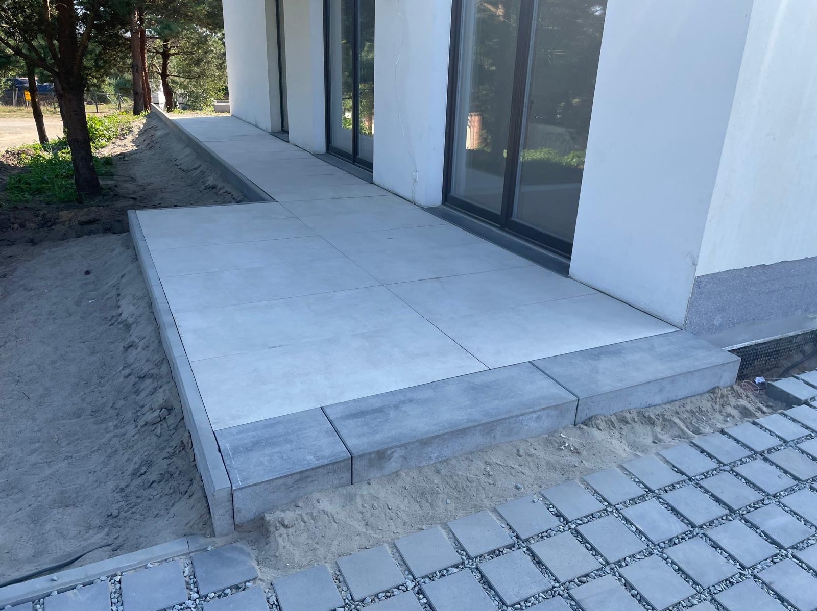 Nowoczesny taras z jasnoszarych płyt, wykończony ciemniejszymi obrzeżami, przylegający do budynku z dużymi oknami. Widoczne przygotowane podłoże z piasku i częściowo ułożona kostka.