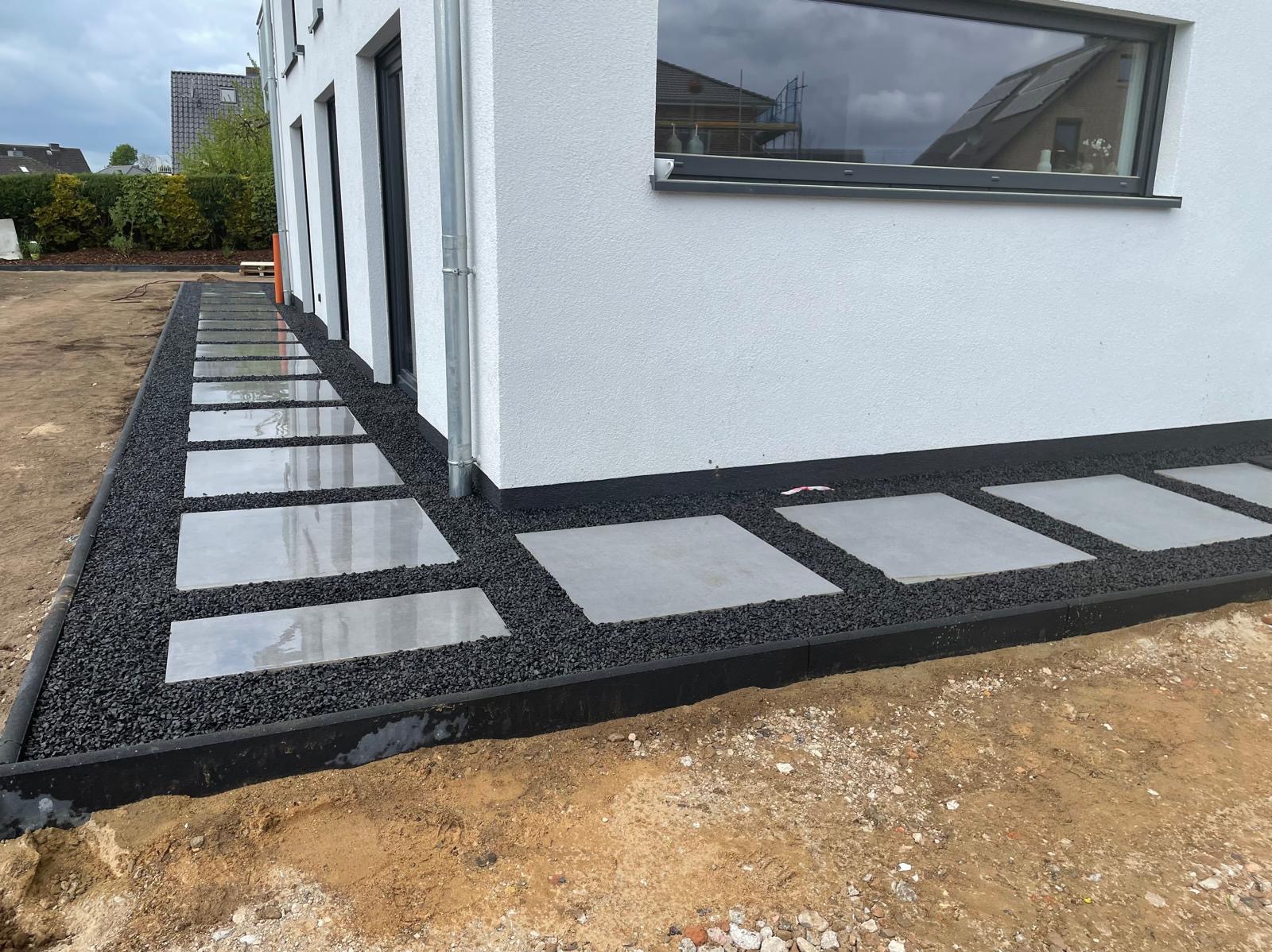 Nowoczesna ścieżka z kwadratowych płyt ułożonych w czarnym żwirze przy białym budynku z dużym oknem. Minimalistyczny design ogrodu.