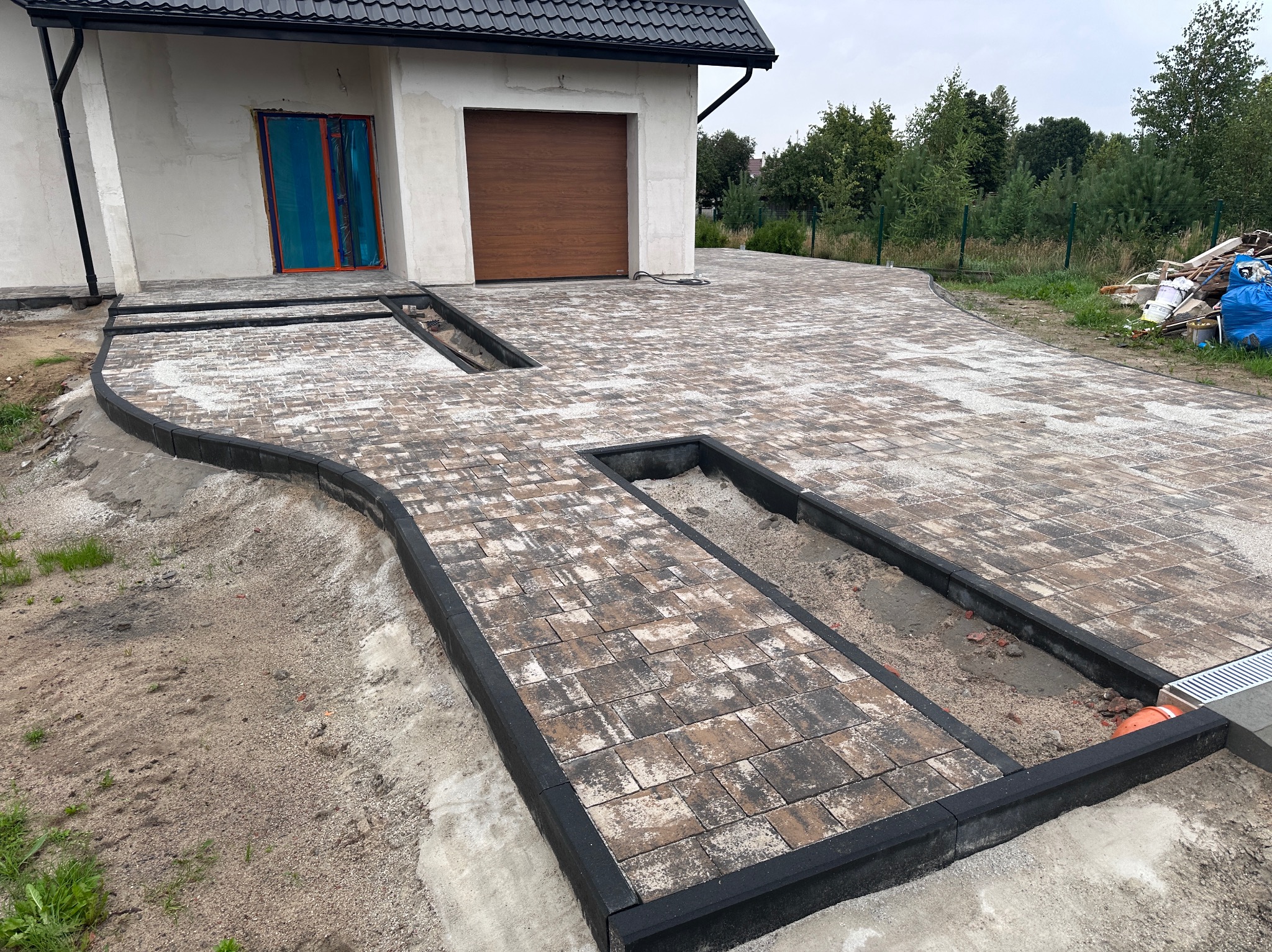 Ułożona kostka brukowa w odcieniach brązu i szarości tworzy ścieżkę i podjazd przed domem z brązową bramą garażową. Widoczne czarne obrzeża i fragmenty niezakończonych prac.