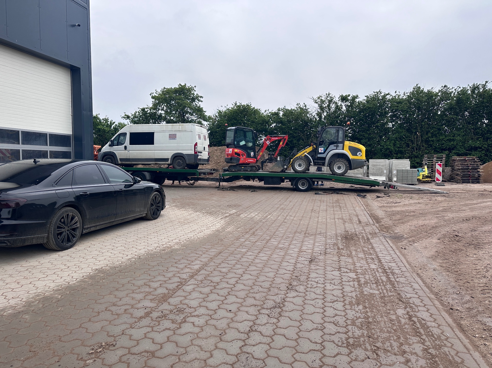 Transport maszyn budowlanych: biały bus, czerwona koparka i żółta ładowarka na niskopodwoziowej przyczepie, zaparkowane na kostce brukowej obok czarnego samochodu.