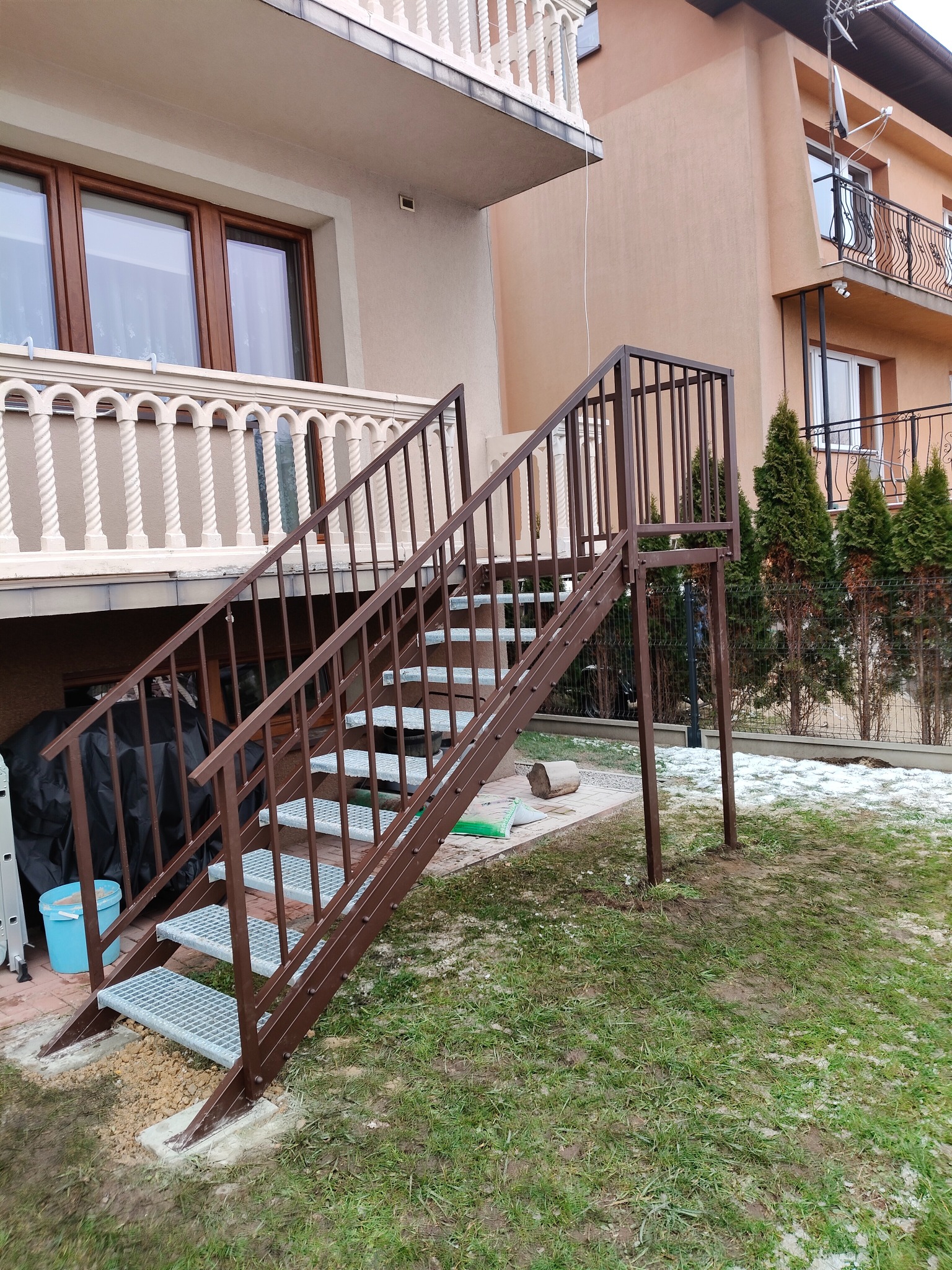 Zewnętrzne schody metalowe w kolorze brązowym, prowadzące z ogrodu na balkon. Widoczne elementy konstrukcyjne i ażurowe stopnie. Tło stanowi budynek z balkonem i fragment ogrodu.