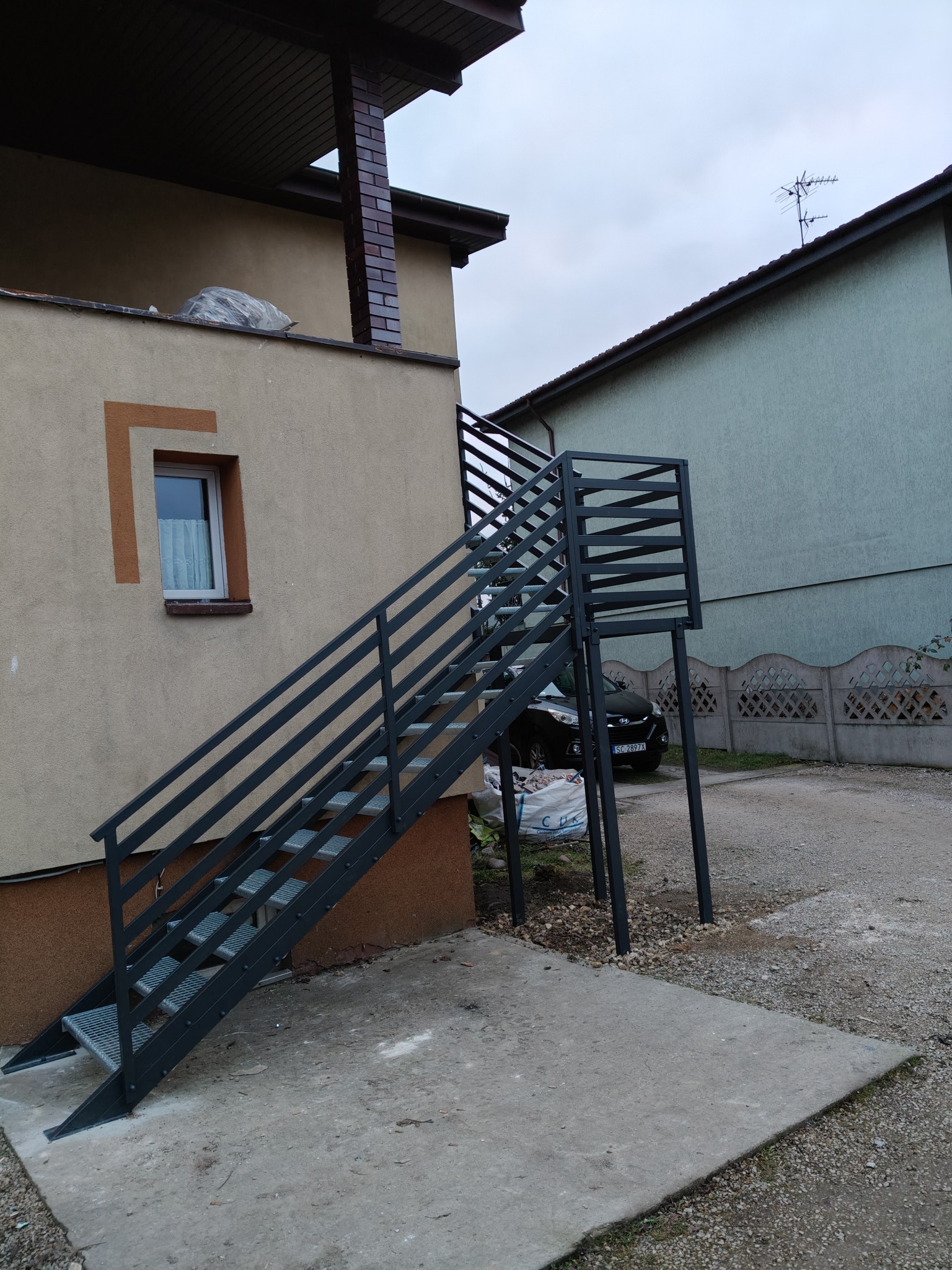 Zewnętrzne schody metalowe z ażurową balustradą, prowadzące do wejścia na piętro budynku. Widoczne elementy konstrukcyjne i otoczenie domu.