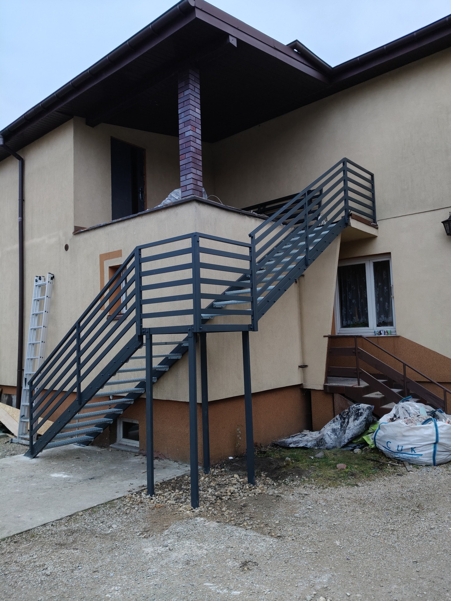 Zewnętrzne schody metalowe z ażurową balustradą prowadzące na balkon domu. Szare, poziome elementy balustrady. Widoczna drabina oparta o ścianę budynku.