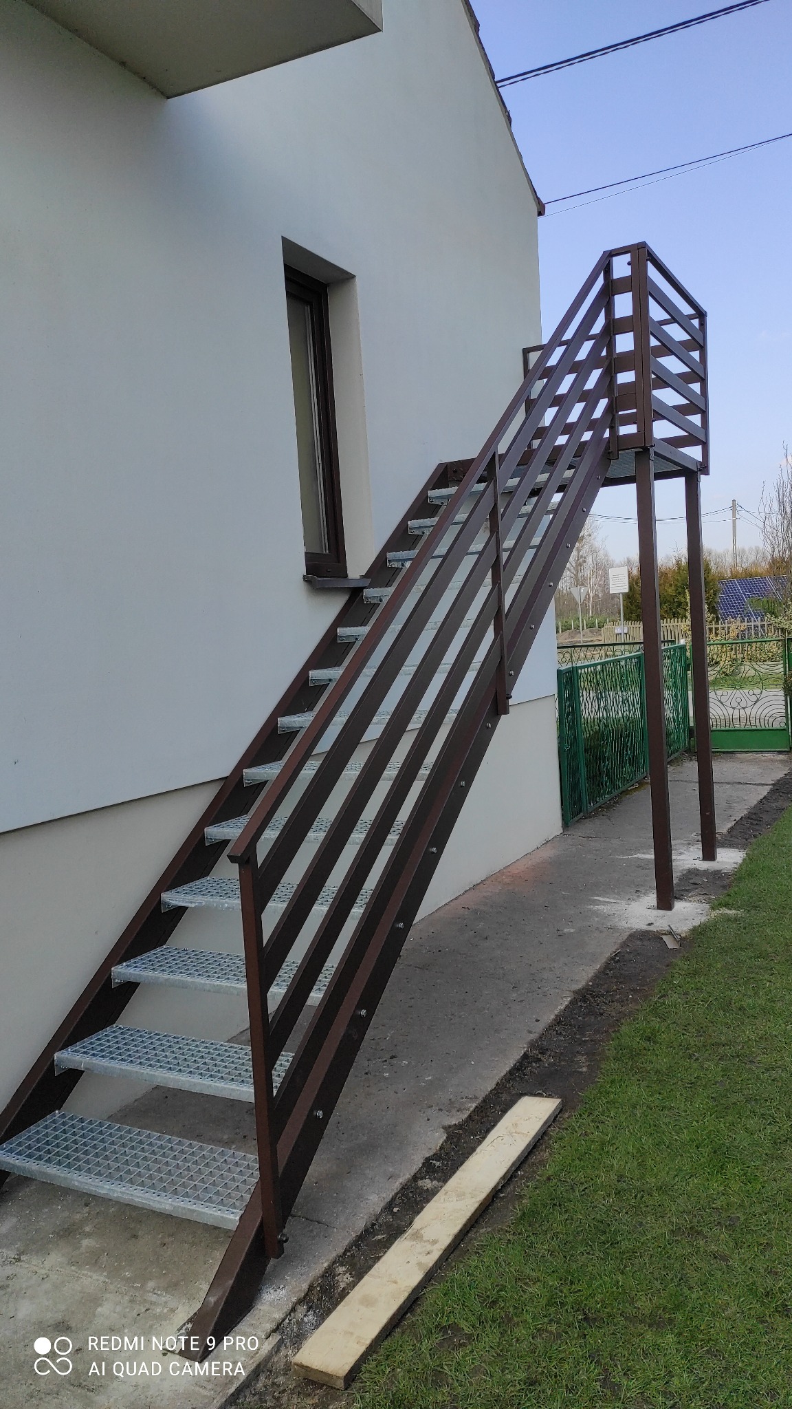 Zewnętrzne schody metalowe z podestem, prowadzące do drzwi na piętrze. Konstrukcja w kolorze brązowym, stopnie z kraty pomostowej. Widok z boku, dom z białego tynku.