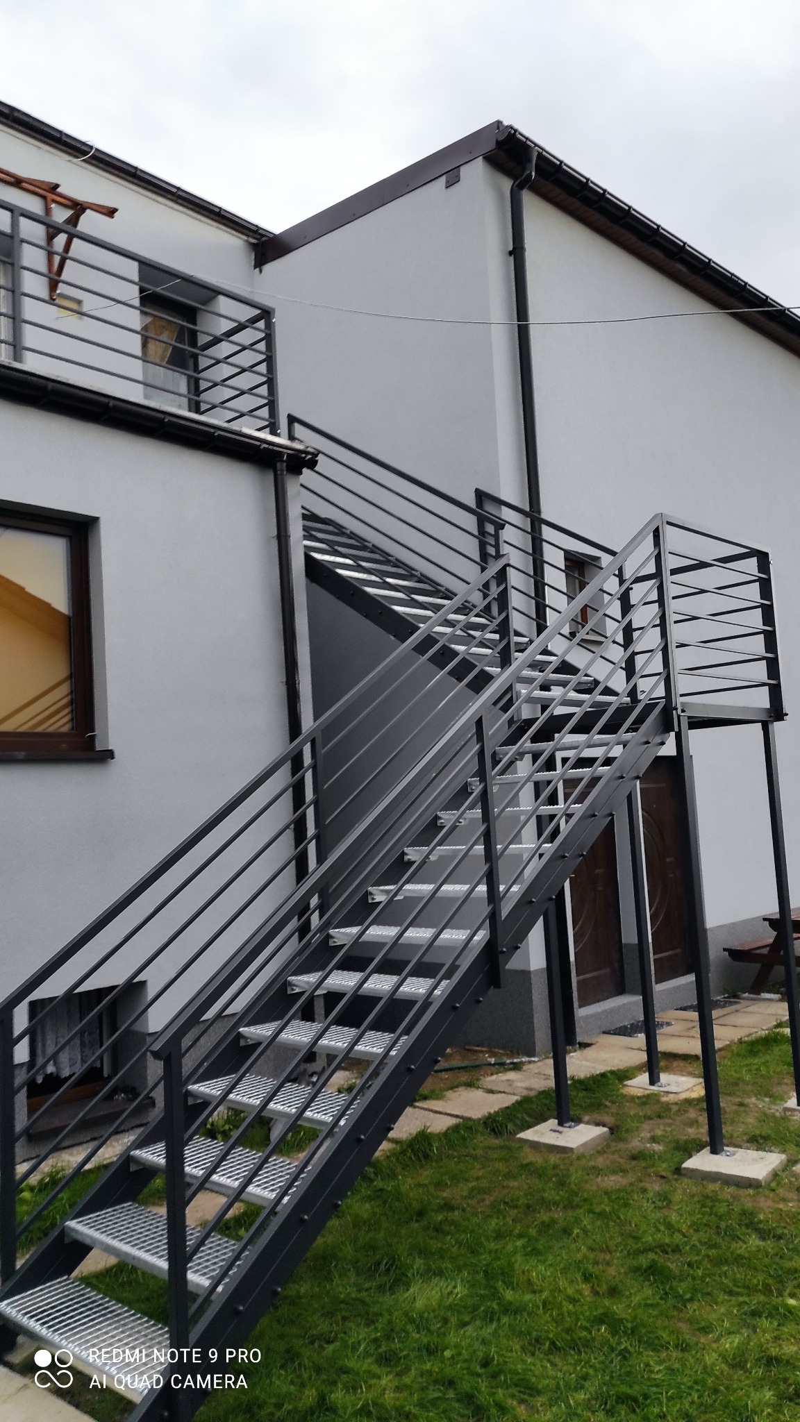 Zewnętrzne schody metalowe z balustradą, prowadzące do drzwi na piętrze budynku. Konstrukcja pomalowana na czarno, stopnie z metalowej kraty. Widok z dołu, lekko z boku.