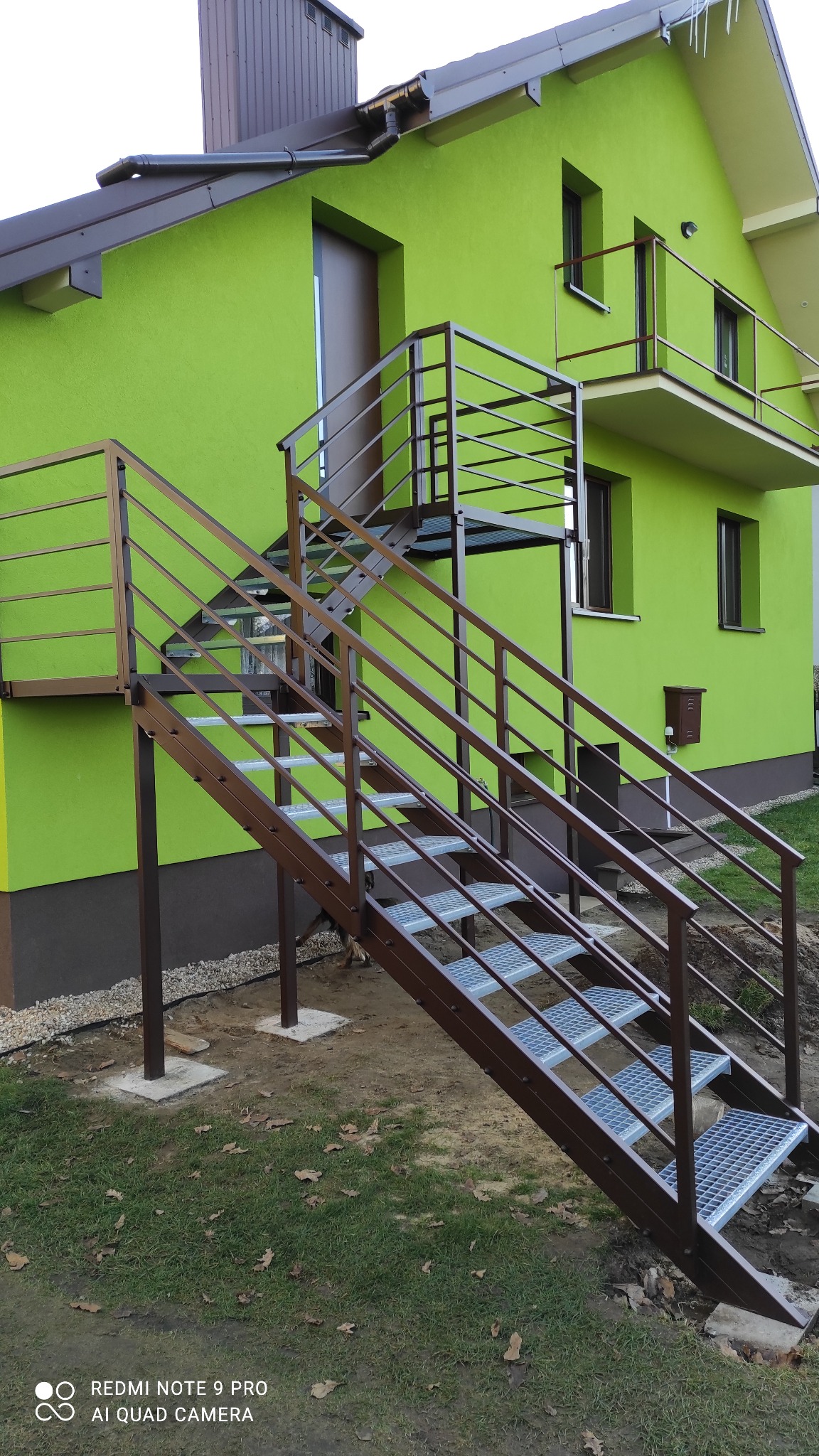 Zewnętrzne schody metalowe z brązowymi poręczami prowadzące do drzwi na piętrze, na tle zielonego domu z balkonem. Widoczne metalowe stopnie i konstrukcja.