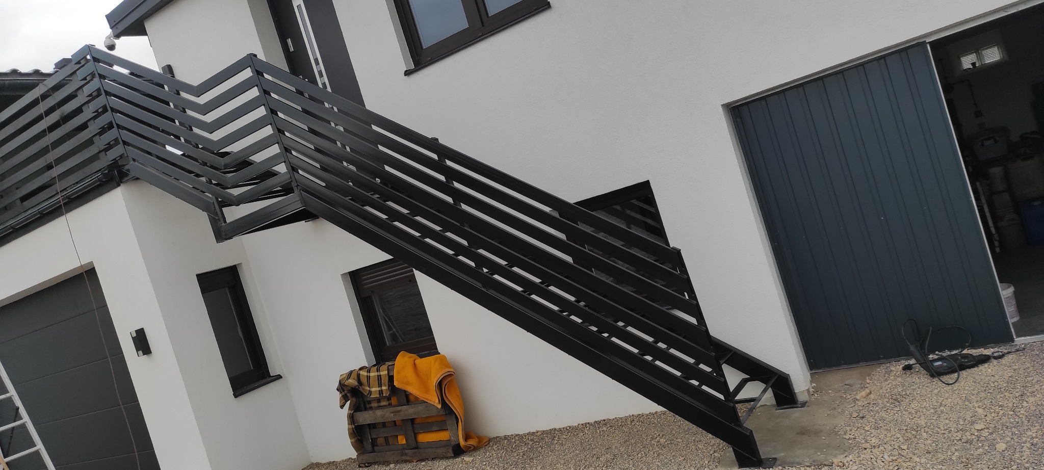 Nowoczesne schody zewnętrzne z czarną, metalową balustradą o geometrycznym wzorze, prowadzące na balkon. Biała elewacja budynku, szara brama garażowa.