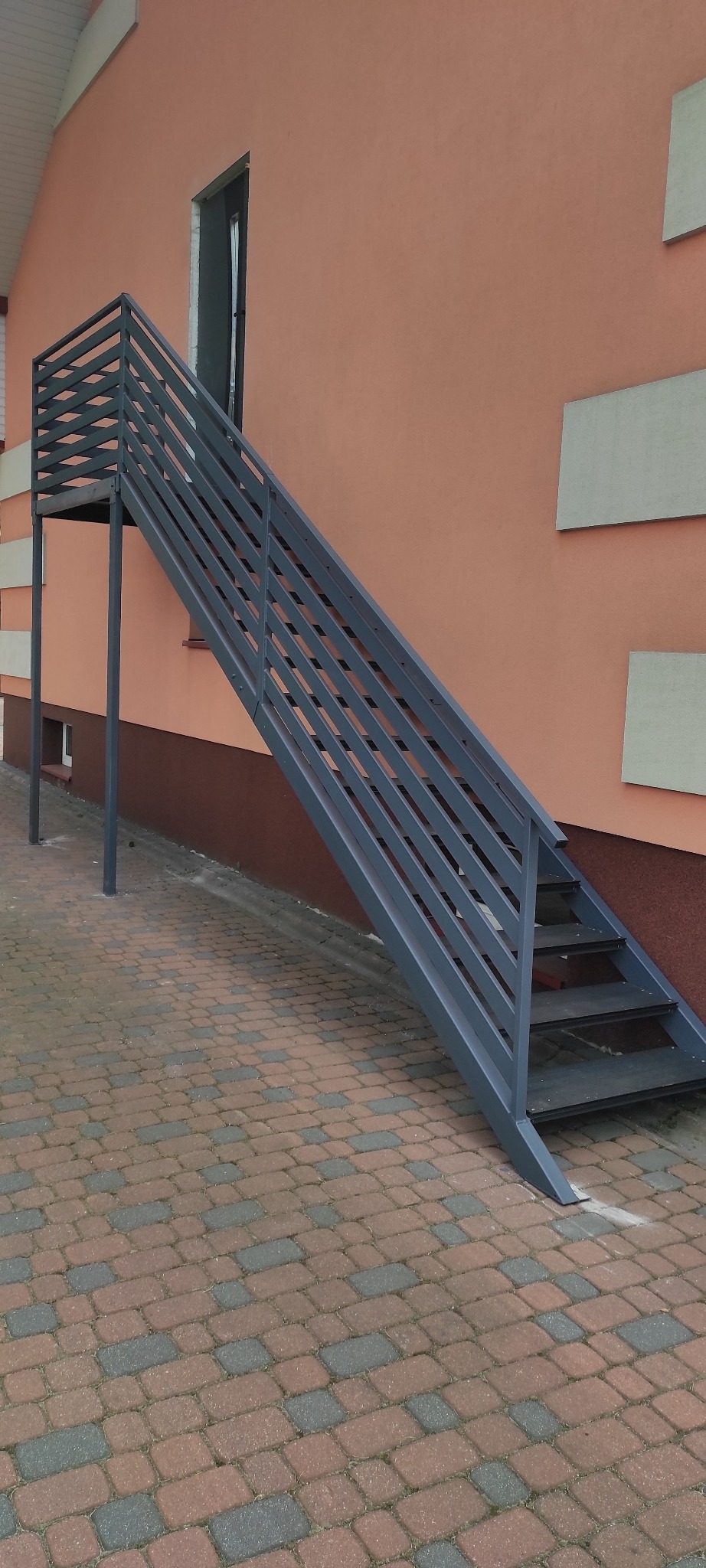 Zewnętrzne schody metalowe z poręczą, prowadzące do drzwi na piętrze. Szare, poziome elementy konstrukcyjne na tle brzoskwiniowej ściany budynku.