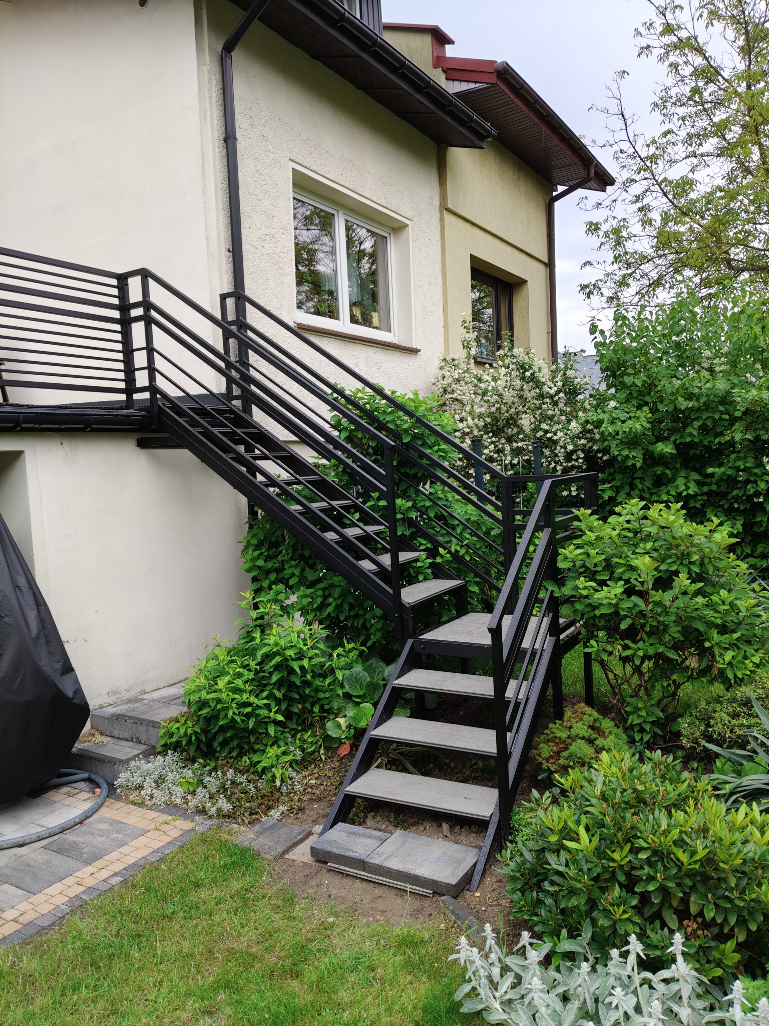 Zewnętrzne schody metalowe z czarną, minimalistyczną konstrukcją, prowadzące do wejścia na piętro budynku, otoczone zielenią ogrodu. Stopnie z szarego materiału.