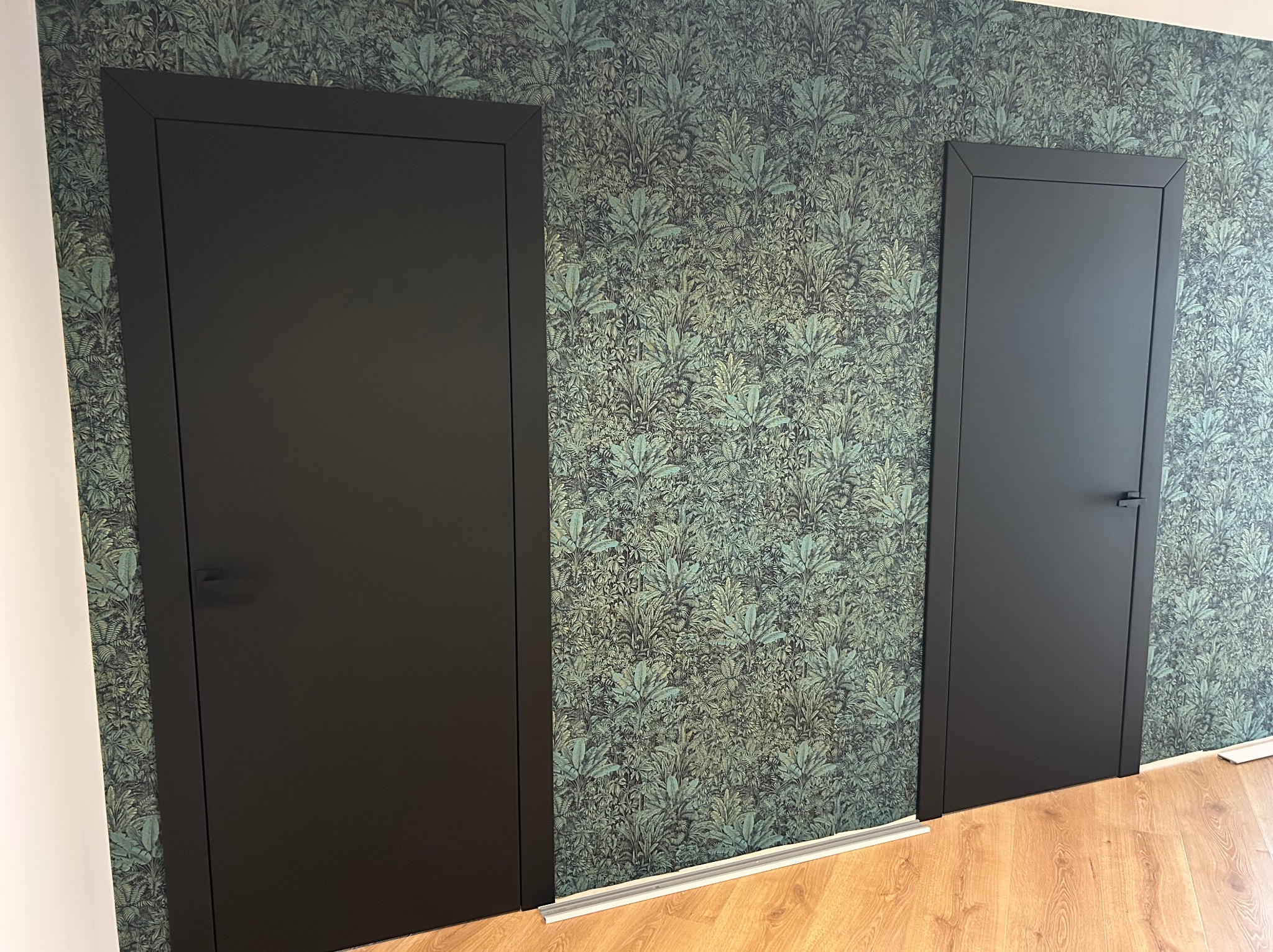 Dwa czarne, minimalistyczne drzwi wewnętrzne na tle ściany w tropikalne wzory. Podłoga drewniana. Ujęcie z bliska, prezentujące detale wykończenia.