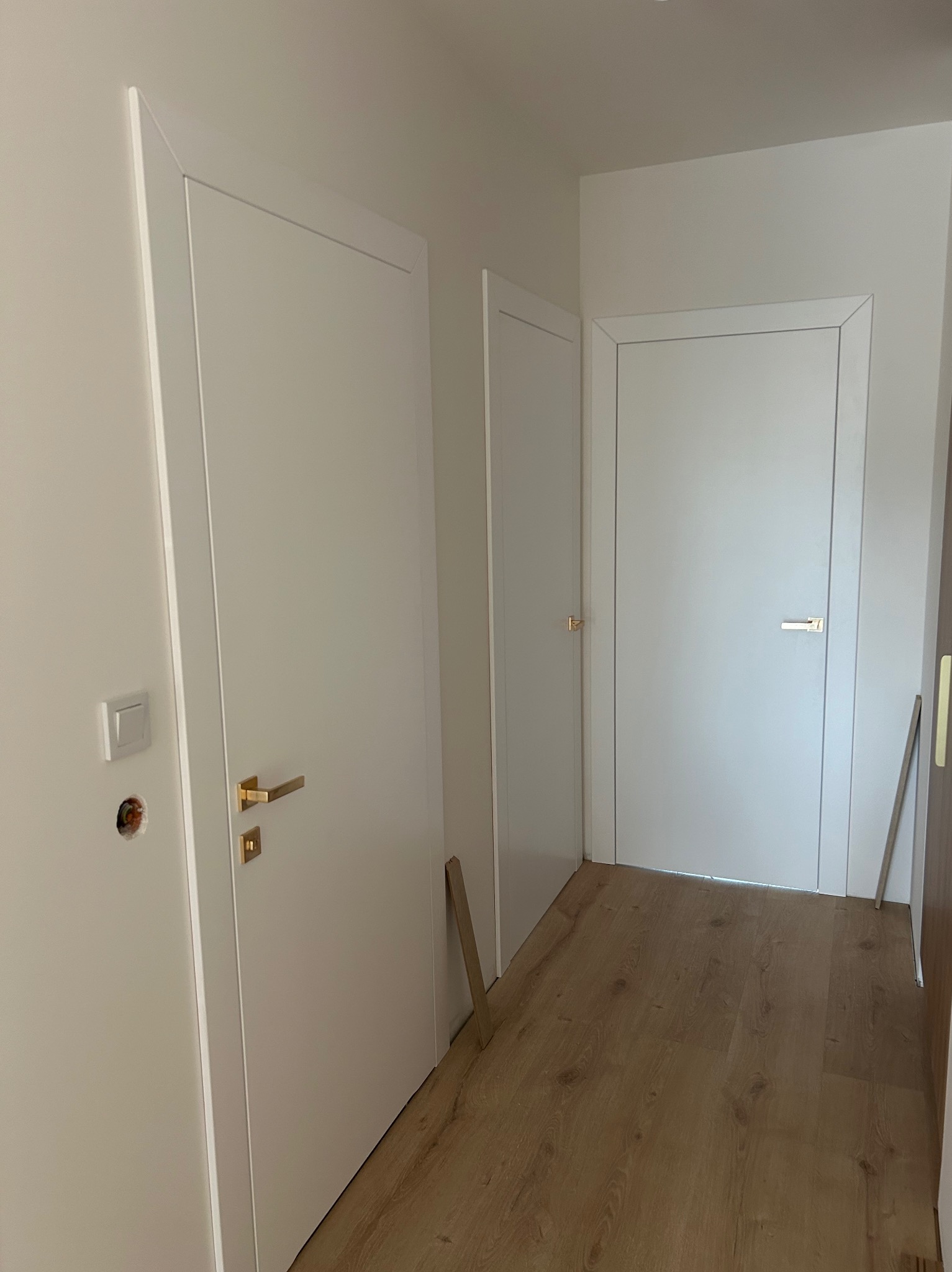 Białe drzwi wewnętrzne z złotymi klamkami w korytarzu, widoczne gniazdko elektryczne bez osprzętu, Skrzydlna. Minimalistyczny design, jasne wnętrze.