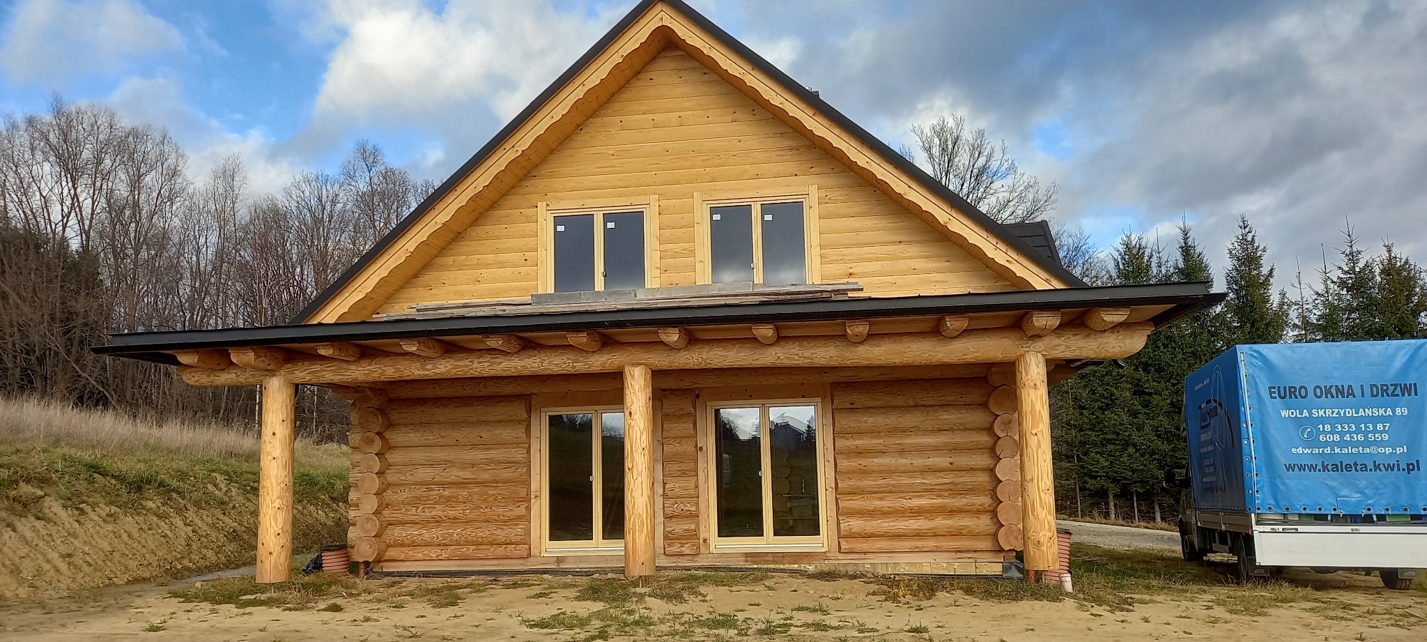 Dom z bali z jasnymi oknami i drzwiami, widoczny fragment niebieskiej plandeki z napisem 'EURO OKNA I DRZWI'.