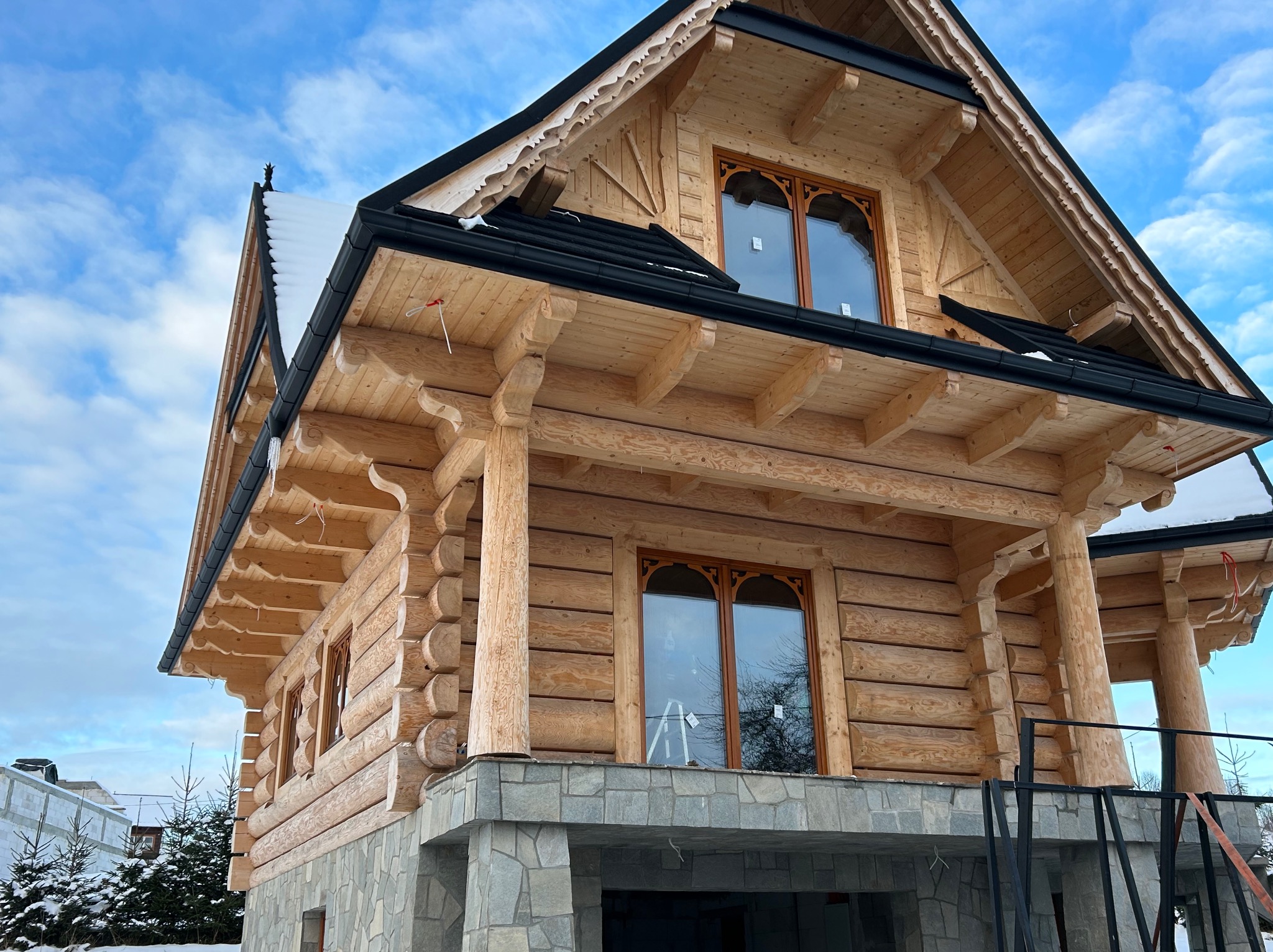 Dwukondygnacyjny dom z bali, z kamienną podmurówką i oknami z dekoracyjnymi łukami, na tle błękitnego nieba. Widoczne detale wykończenia dachu i drewnianych elementów konstrukcyjnych.