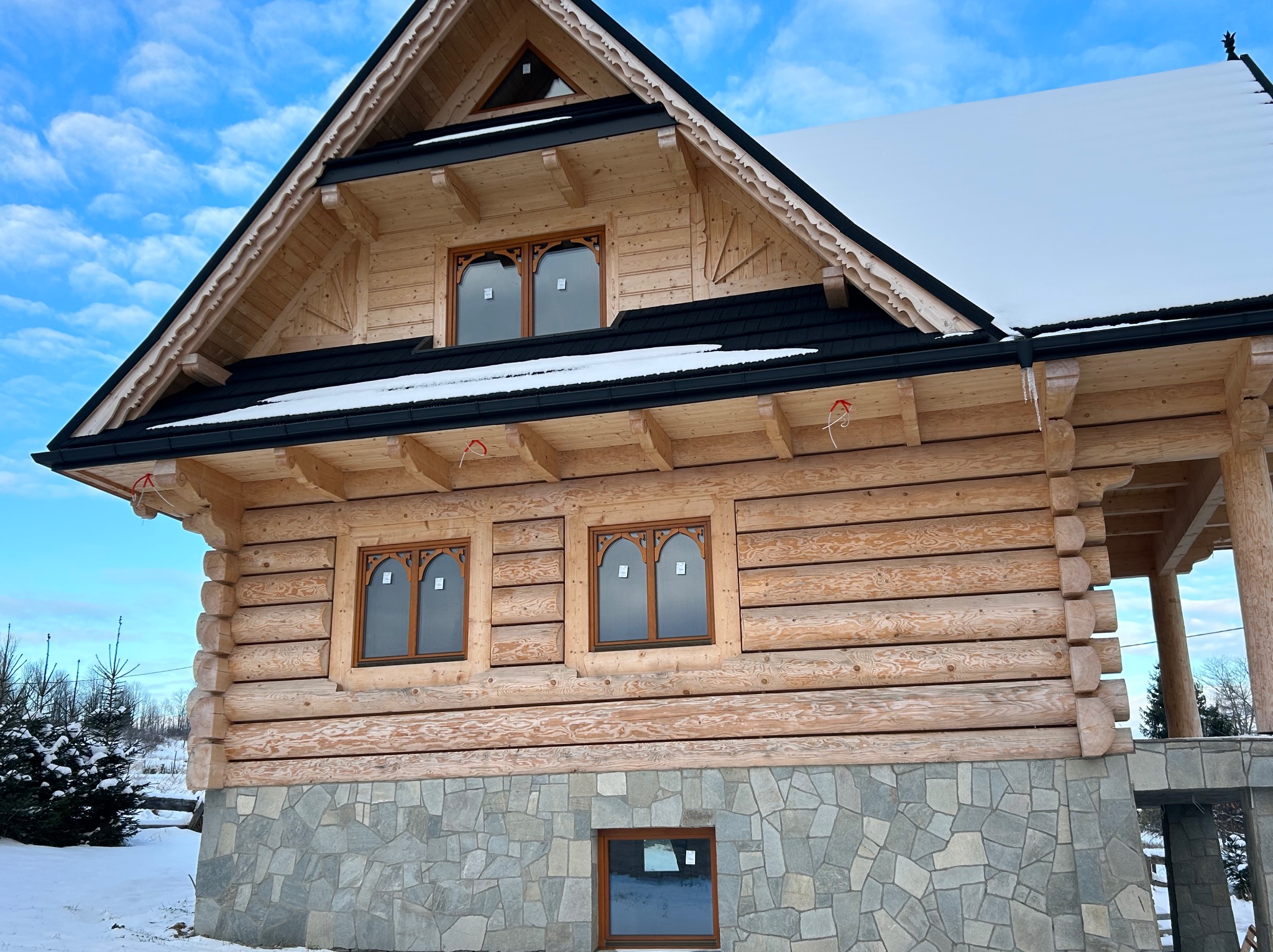 Drewniany dom w stylu góralskim z kamienną podmurówką i oknami z ozdobnymi szprosami, częściowo ośnieżony, na tle błękitnego nieba.