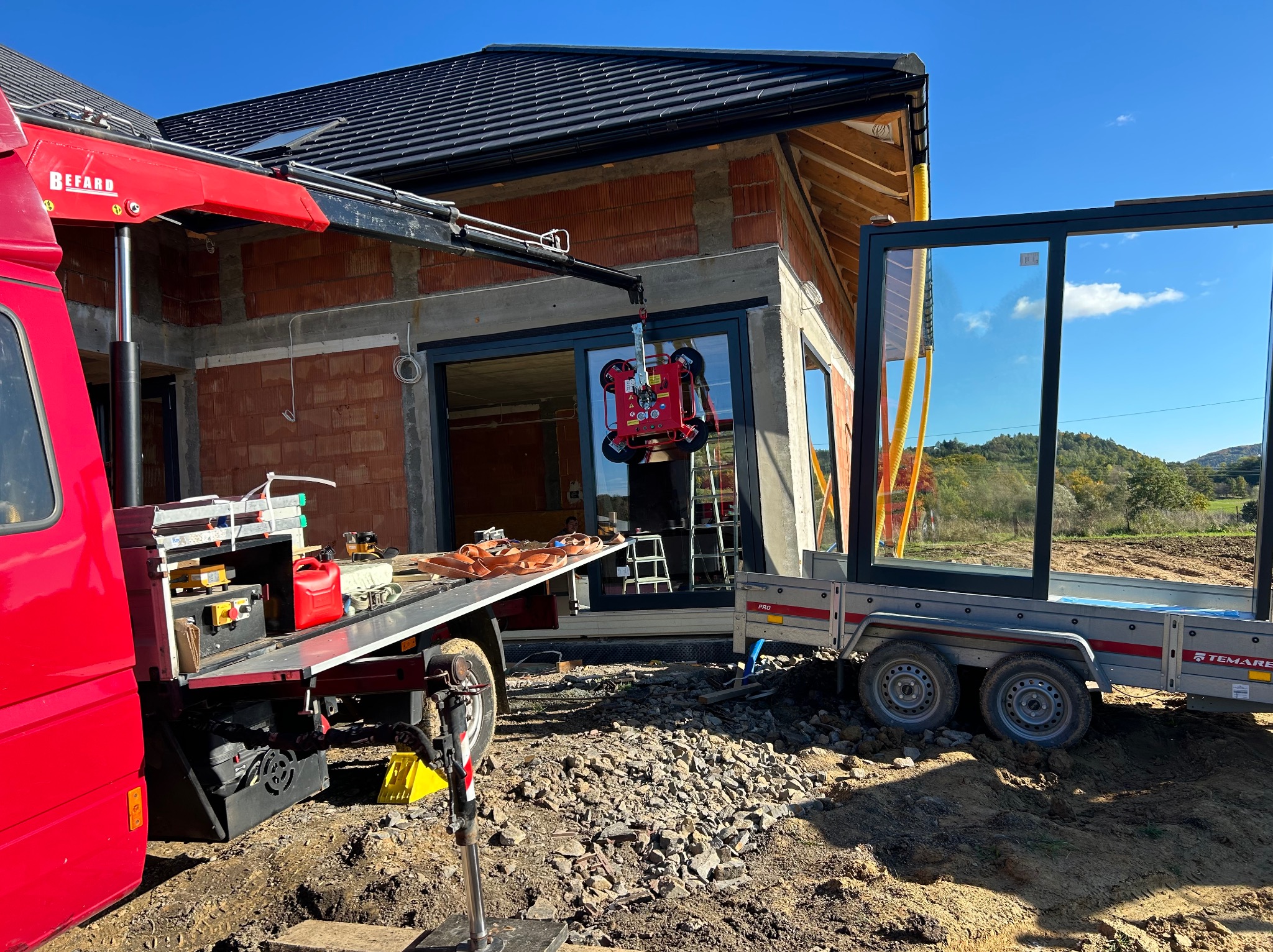 Montaż okna w surowym budynku z użyciem przyssawki i dźwigu. Okno na przyczepie obok. Widoczne narzędzia i gruz na placu budowy. Słoneczny dzień.