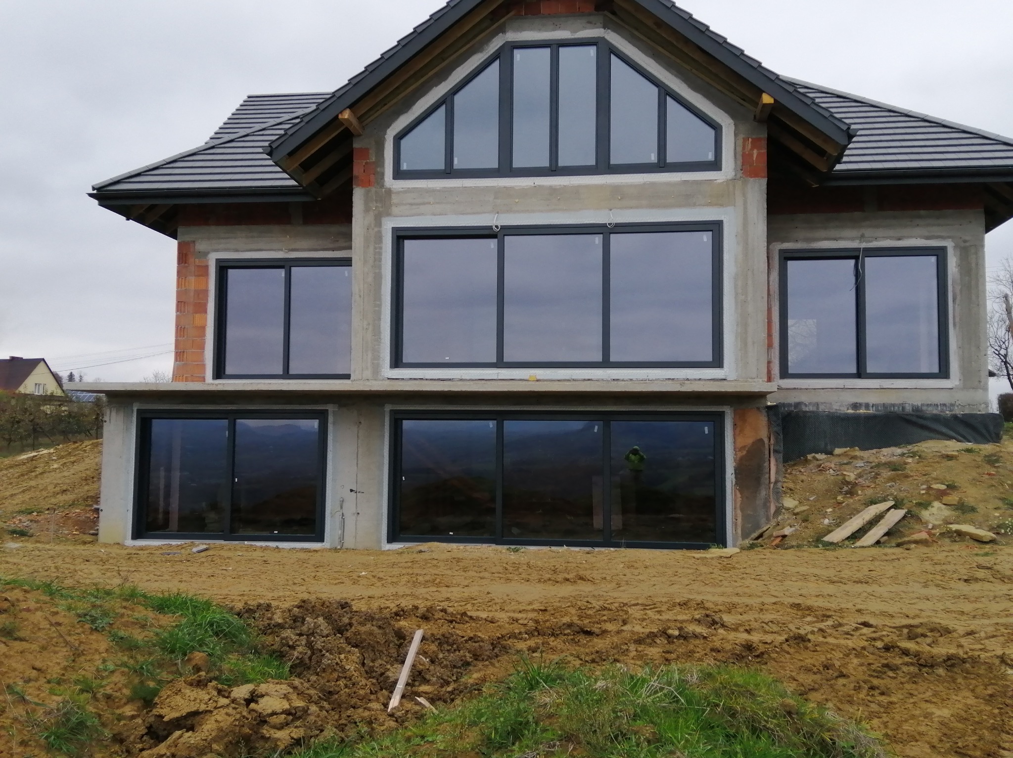 Surowy stan domu z dużymi, nowoczesnymi oknami w ciemnych ramach. Widoczne niedokończone elewacje i ziemia wokół budynku. W oknach odbija się krajobraz.