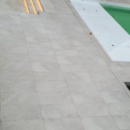 Adrian Durak - Jasne płytki tarasowe ułożone wokół basenu i schodów z oświetleniem LED, widok z góry na dom z ciemną dachówką i drewnianymi elementami wykończeniowymi.