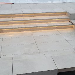 Adrian Durak - Wykończony taras z jasnych płyt z subtelnym oświetleniem schodów. Widoczne detale łączeń płyt i precyzyjne wykonanie. Minimalistyczny design.