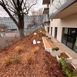 Adrian Durak - Nowoczesny ogród przy budynku z ławką z drewna, rabatami i dekoracyjnymi lampami. W tle budynek szkoły i wieżowiec. Całość utrzymana w stonowanej kolorystyce.